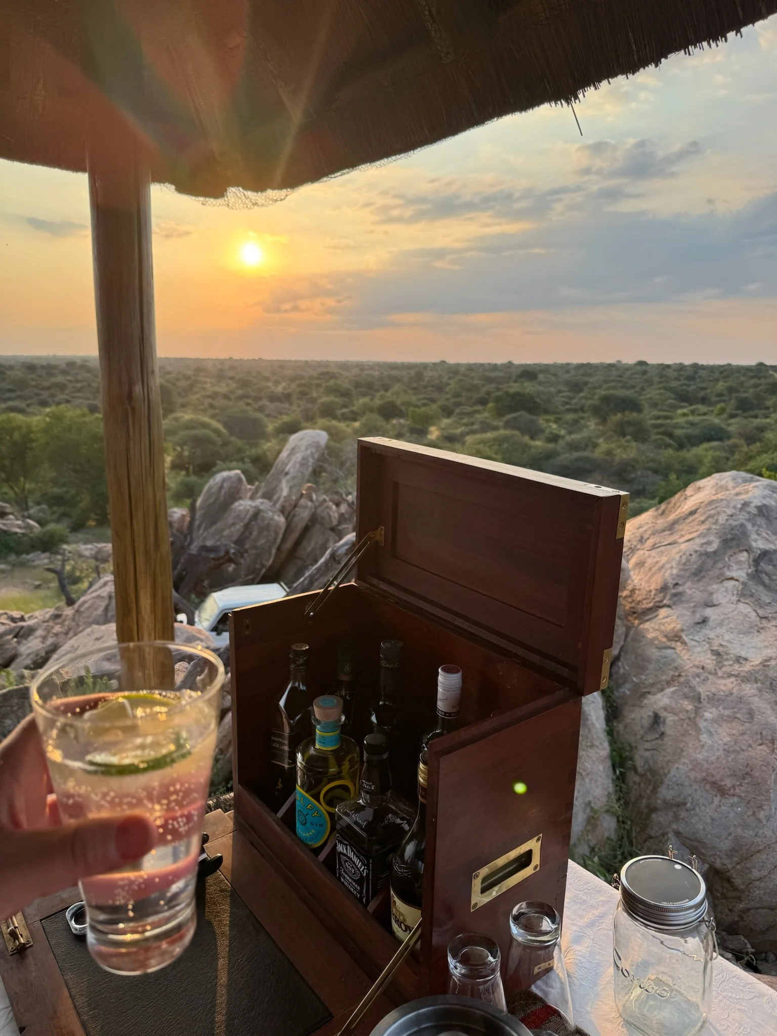 View of a sunset over a vast green landscape with rocks and trees, taken from a shaded outdoor area. In the foreground, a person is holding a glass of clear soda with lime, and there is a wooden box with liquor bottles on a table.