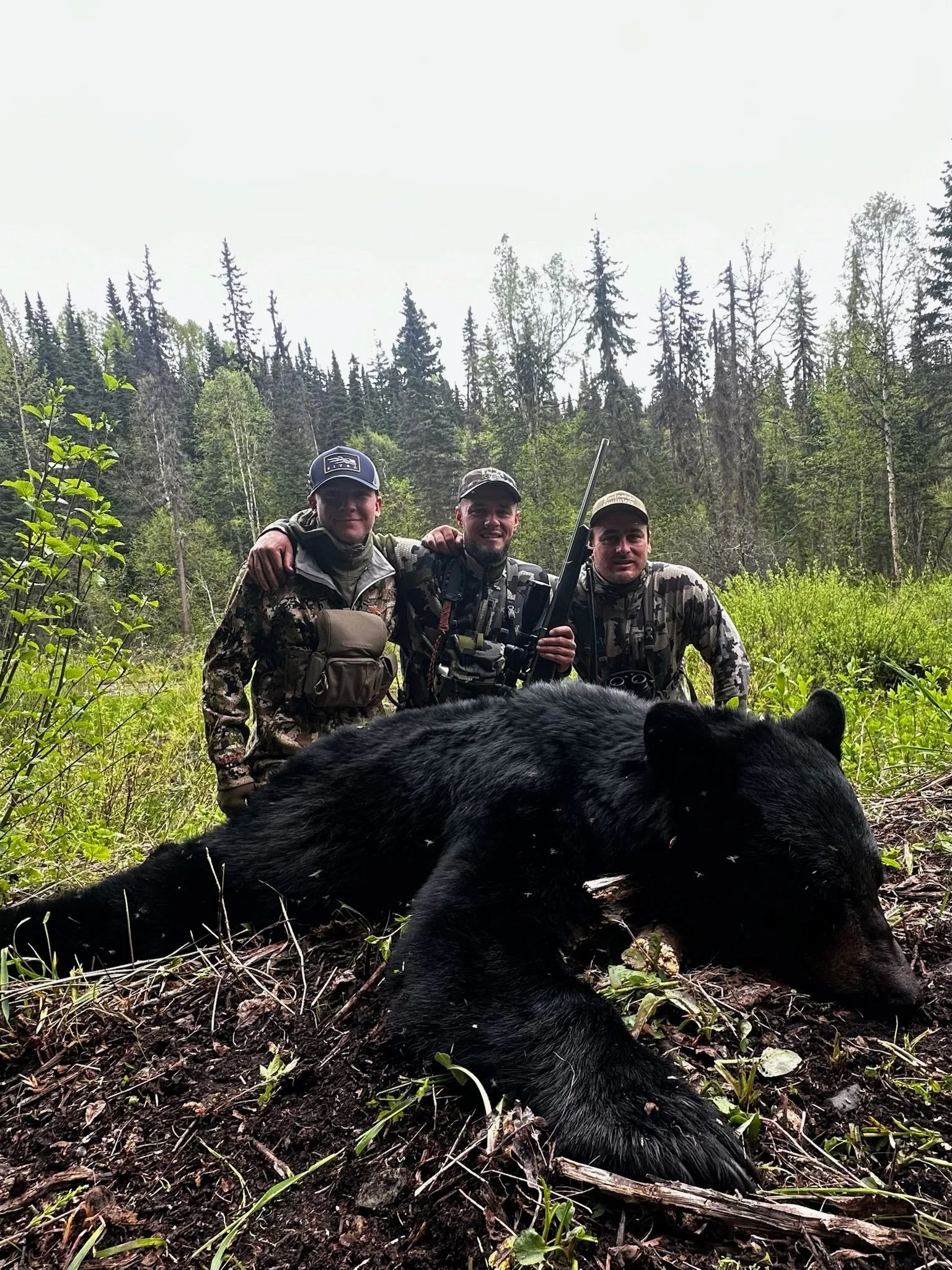Three hunters in camouflage gear pose behind a black bear lying on the ground in a forested area.
