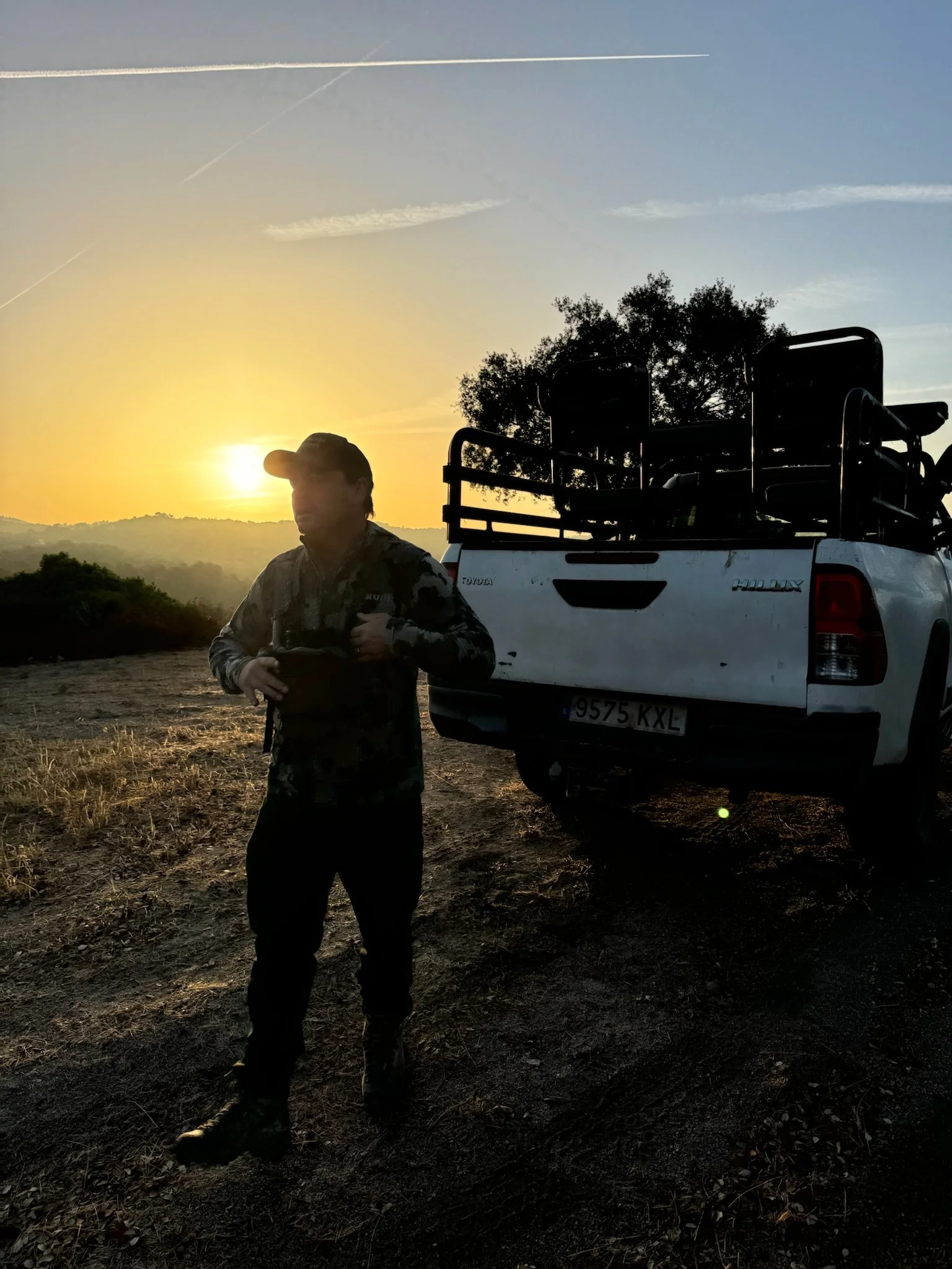 A man in camouflage clothing and a baseball cap stands outdoors near a white pickup truck during sunset. The truck's bed is equipped with a roll cage, and there's a tree and distant hills in the background.