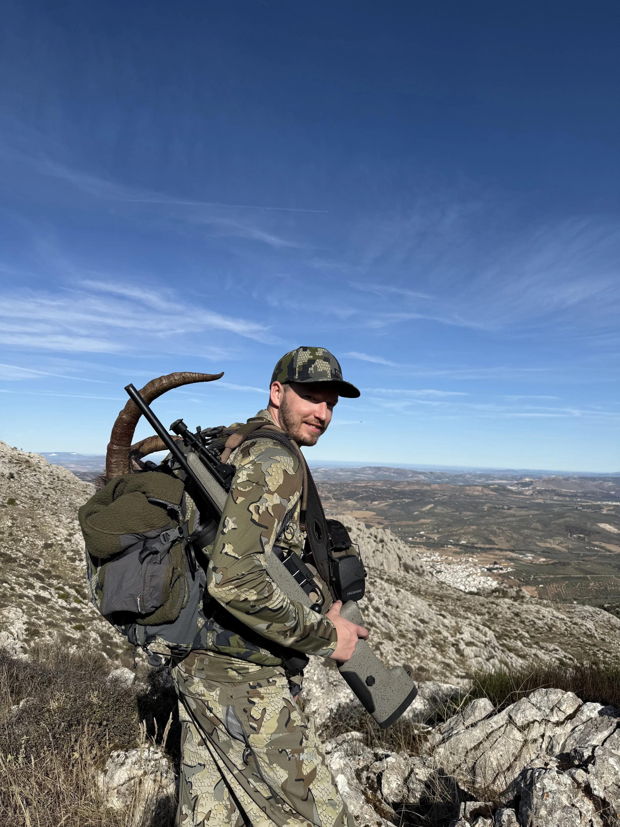 A man dressed in camouflage clothing carrying a large backpack and holding a firearm standing on rocky terrain on a mountain, with a vast landscape of fields and hills in the background under a blue sky.