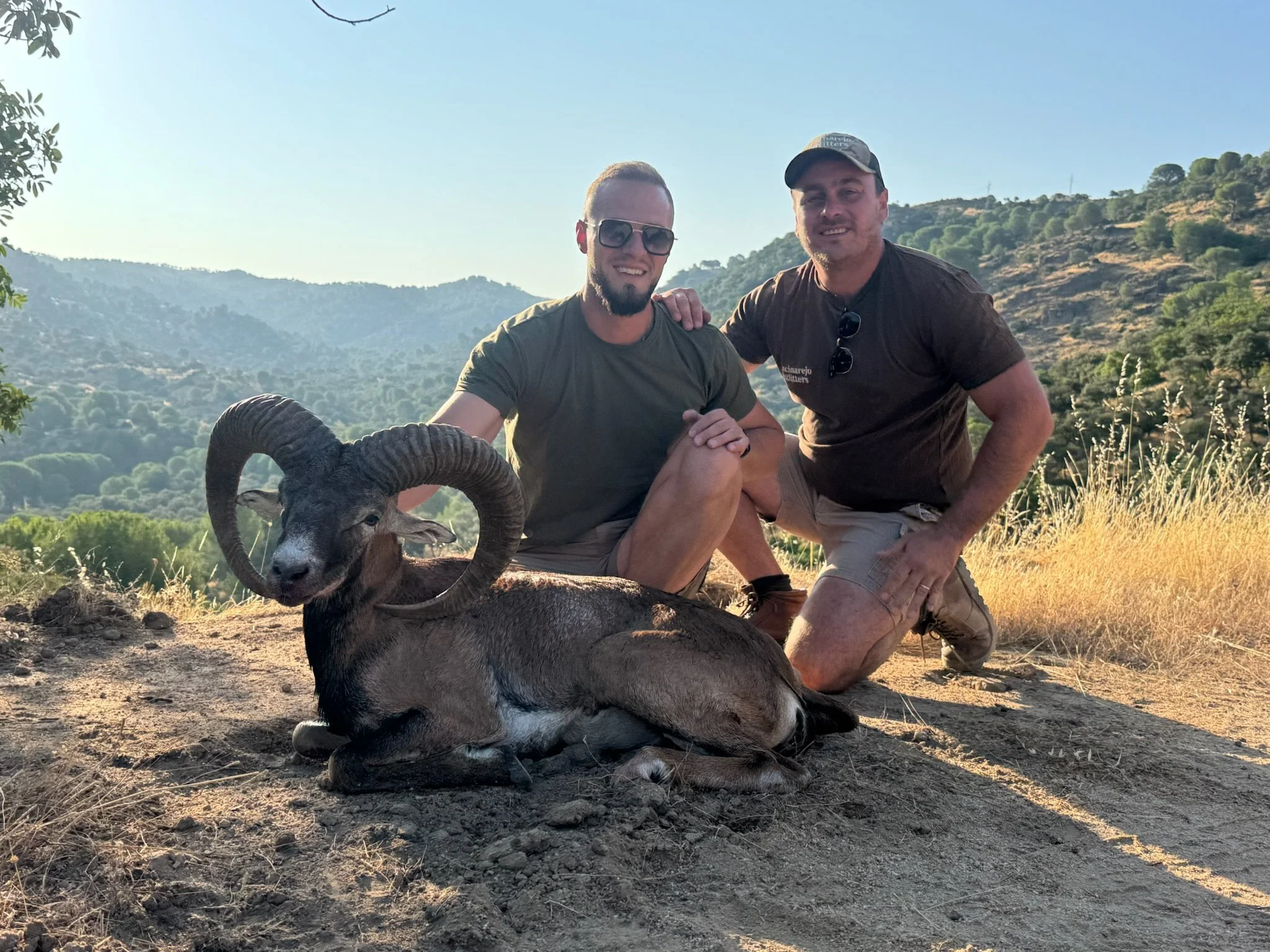 Two men kneel on a dirt patch in a scenic outdoor area with rolling hills and dry grass. One is holding a large ram with prominent curved horns. Both men are smiling and wearing casual outdoor clothing.