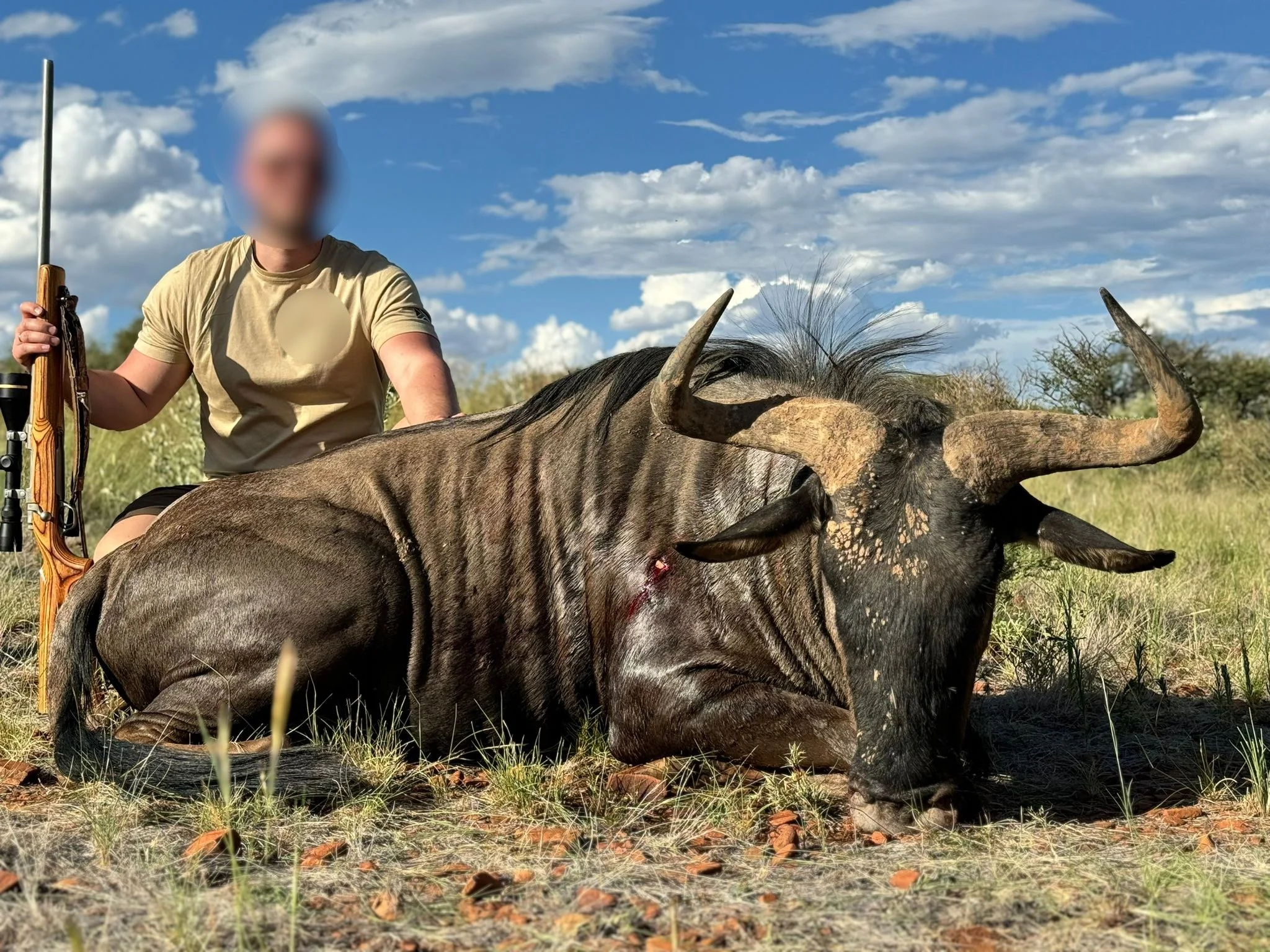 A hunter kneeling next to a large, deceased wildebeest lying on the ground in an open grassy field under a partly cloudy sky.