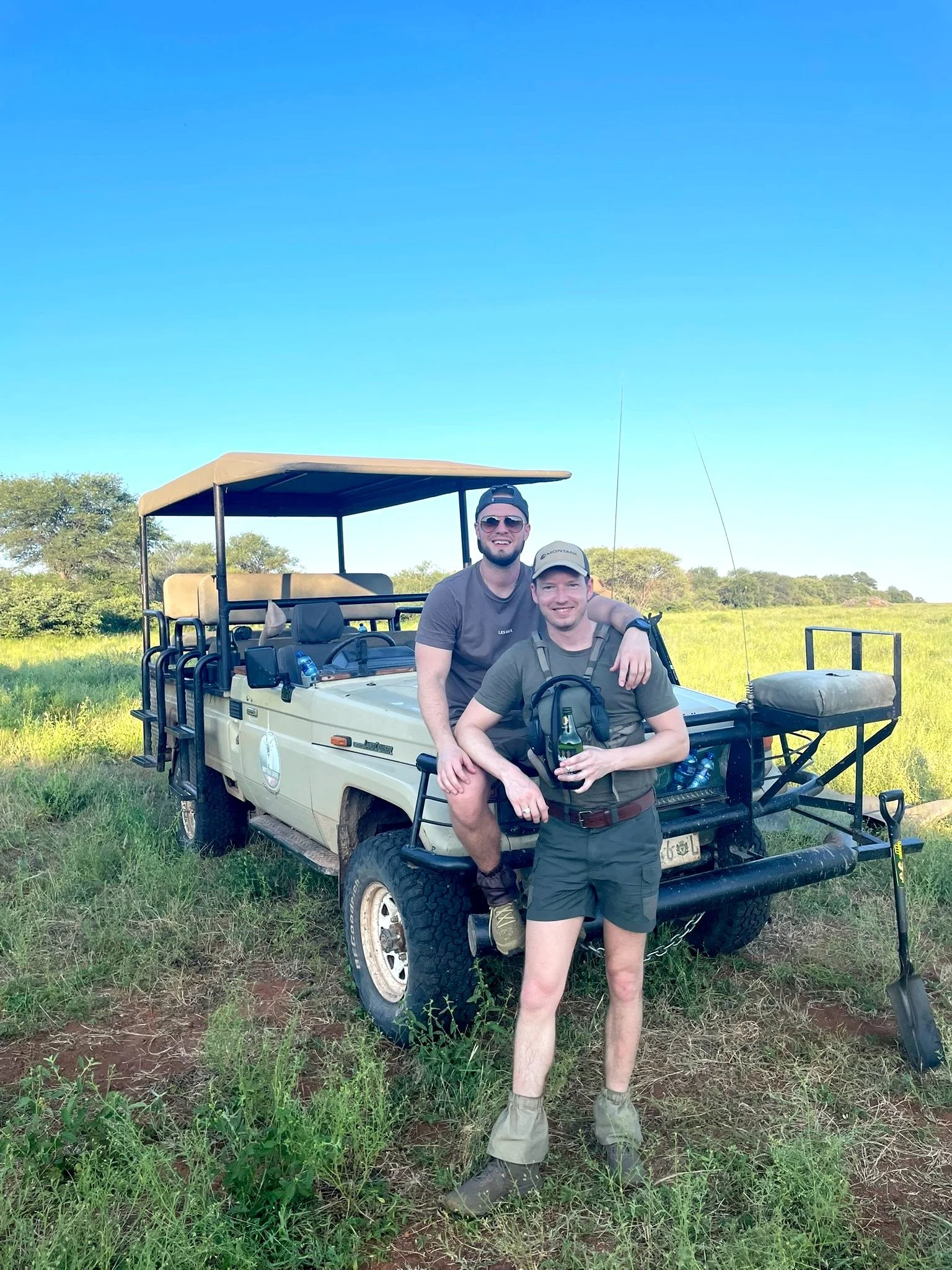 Two men in casual clothing and hats smiling and posing near a safari vehicle in a grassy field, with trees and a clear blue sky in the background.