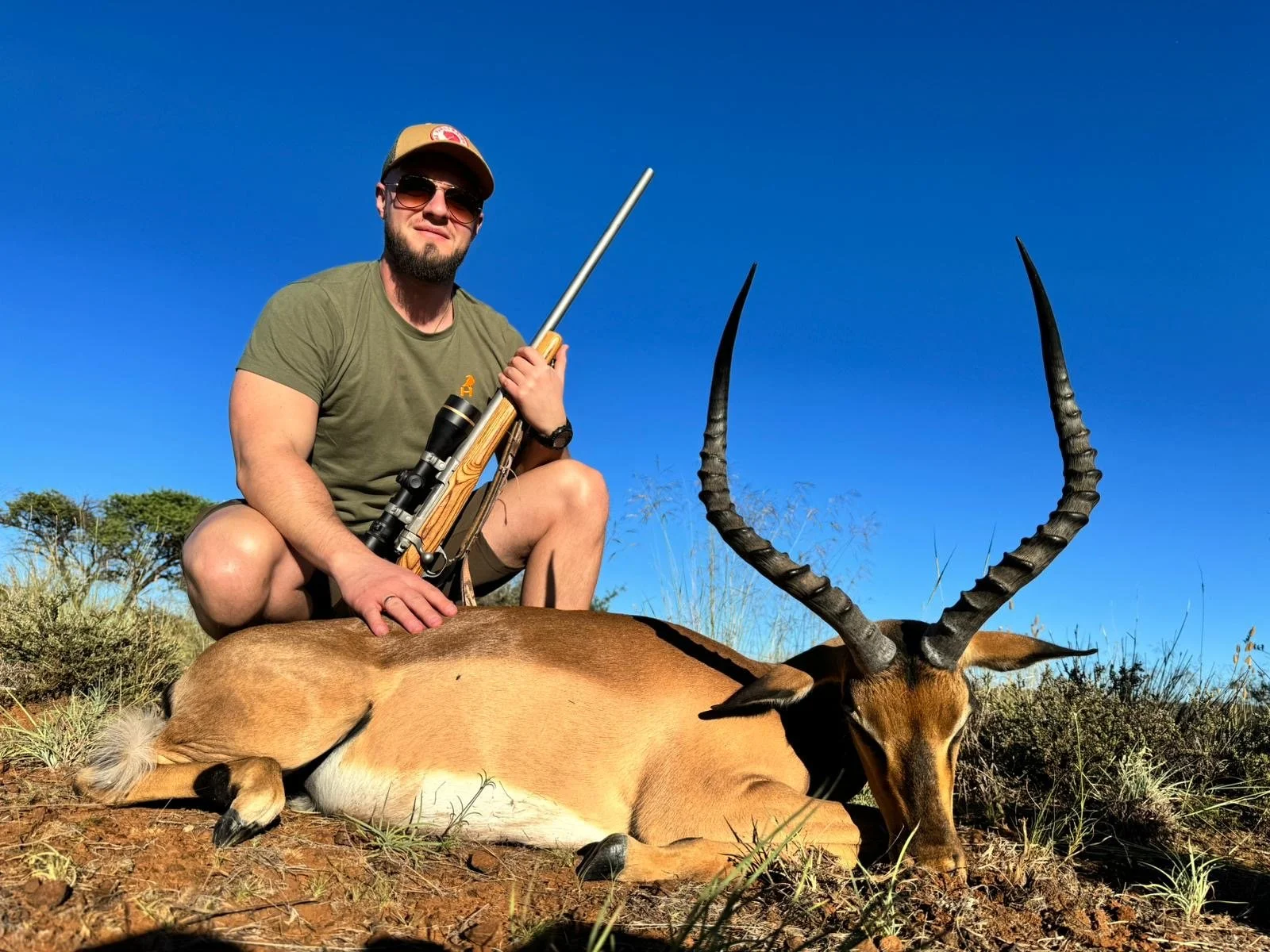Man with sunglasses and a cap kneeling next to a deceased antelope with long curved horns, holding a rifle, in a dry grassy landscape under a clear blue sky.