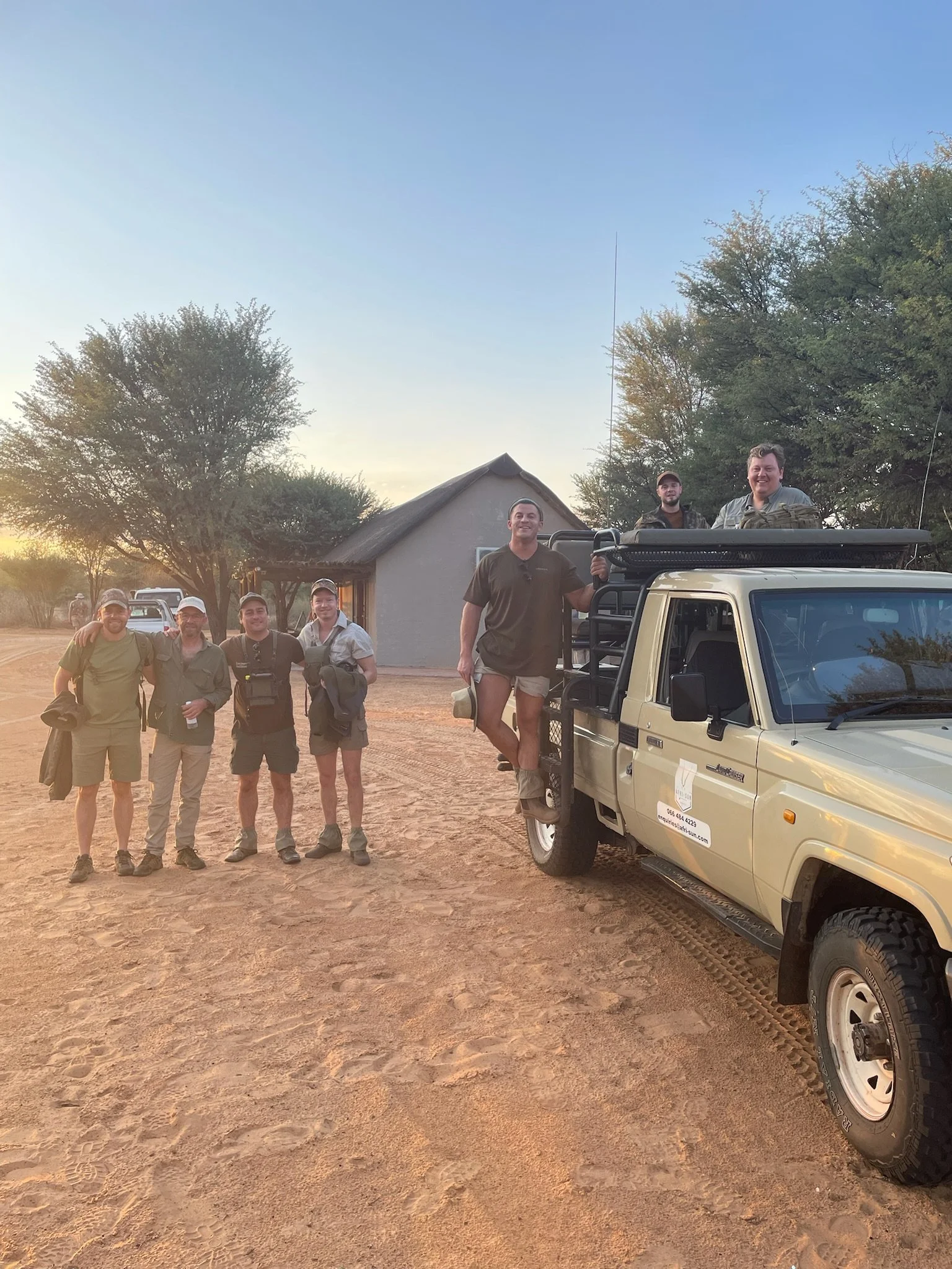 Group of six people standing and sitting on a beige safari vehicle in front of a small house and trees during sunset.