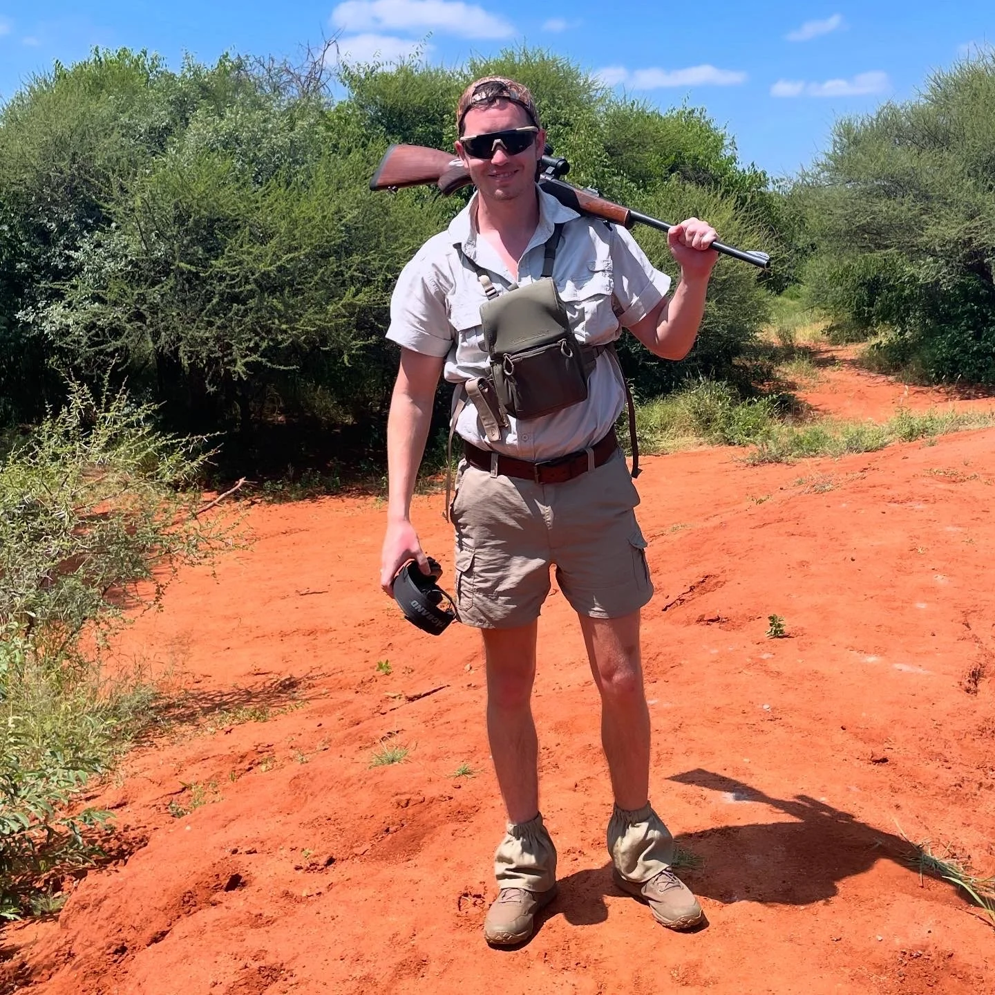 A person standing outdoors on red dirt ground, holding a rifle over their shoulder and wearing sunglasses, a beige shirt, khaki shorts, and hiking boots, with trees and blue sky in the background.