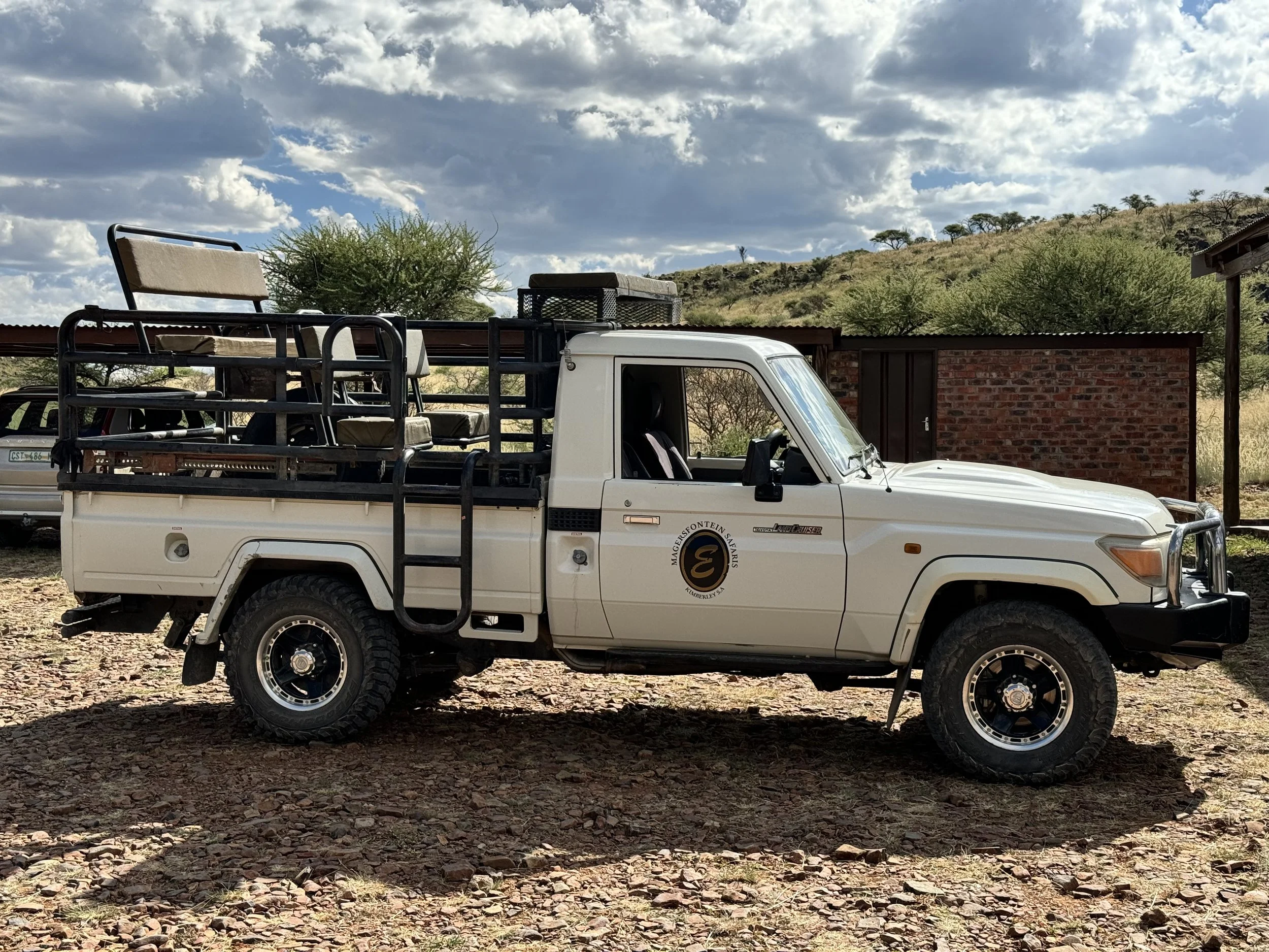 A white utility truck with a logo on the door, parked on a dirt ground with scattered rocks, in an outdoor setting with trees, a brick building, and a cloudy sky in the background.