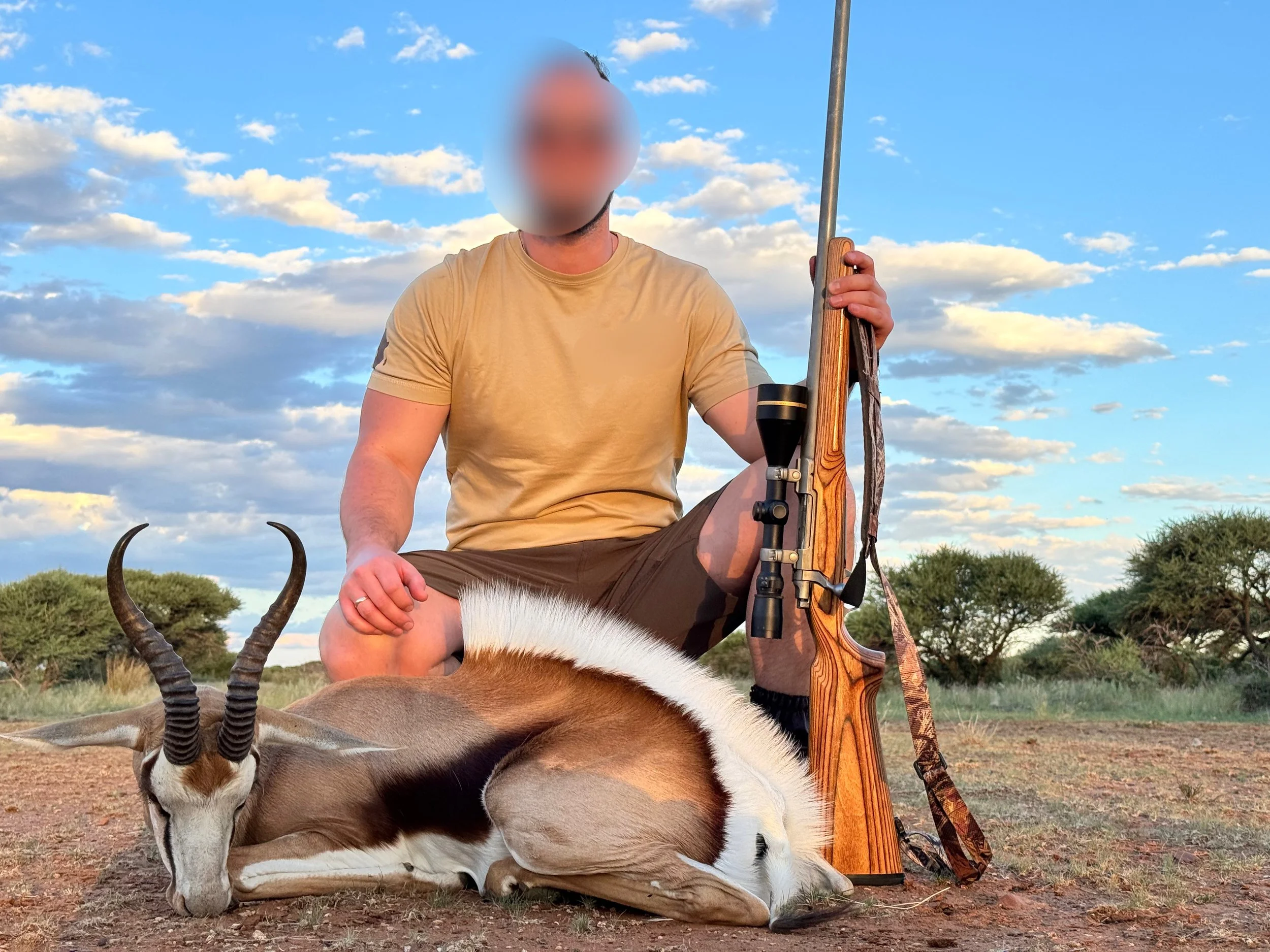 A man kneeling in an open field with a dead antelope and a rifle, with trees and a blue sky with clouds in the background.