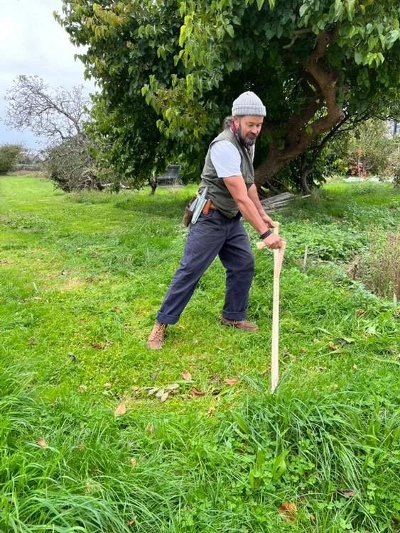Gardener, Danny Parrack, wearing a gray beanie, gray t-shirt, and pants using a sithe on a grassy area as a natural lawncare practise.