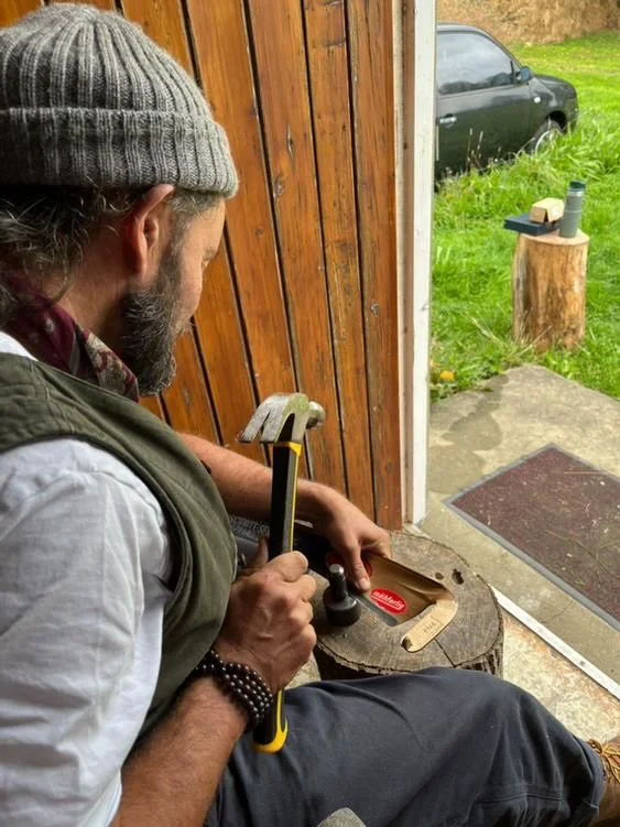 A man wearing a gray beanie, white shirt, and green vest, sitting on a wooden stool and maintaining a sithe tool, with the open door revealing grass and a tree stump outside.