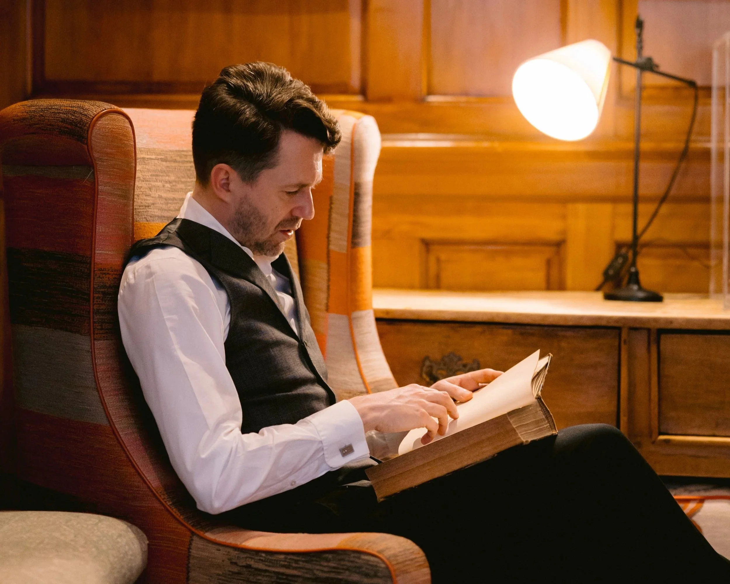 A man with dark hair and facial hair, wearing a white shirt and dark vest, is sitting on a cushioned chair with red and orange striped upholstery. He is reading a newspaper in a room with wood-paneled walls and a large, bright standing lamp.