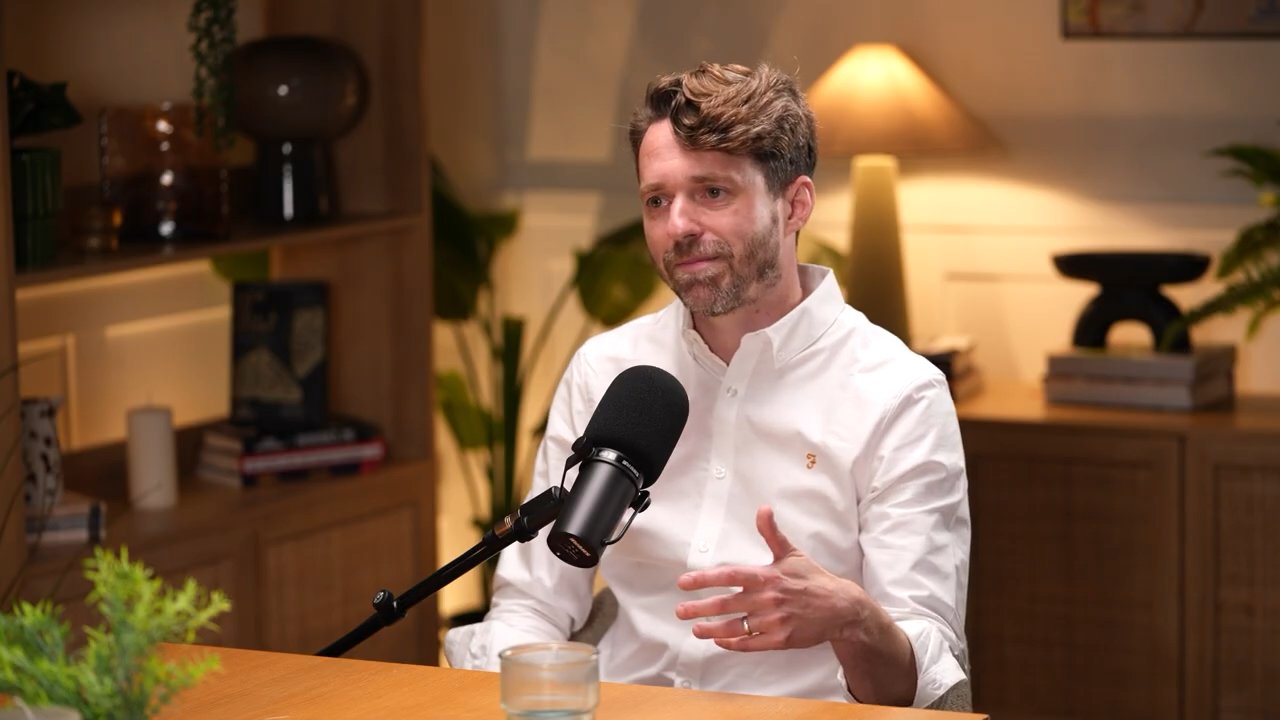 A man in a white shirt sitting in a room with a microphone in front of him, gesturing with his hand, with warm lighting and plants in the background.