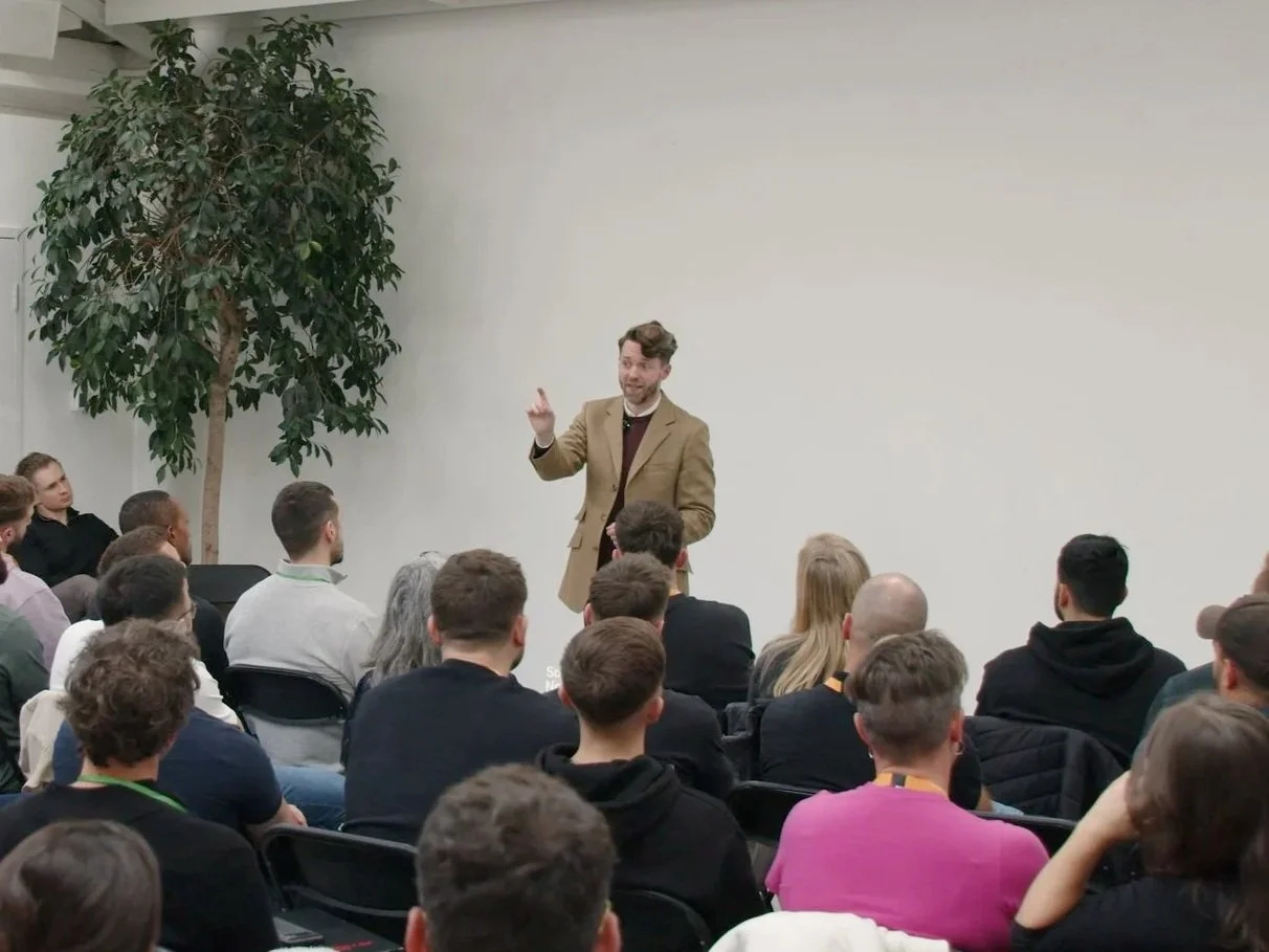 A man in a brown blazer and maroon sweater giving a presentation to an audience in a conference room with a large plant and plain wall behind him.