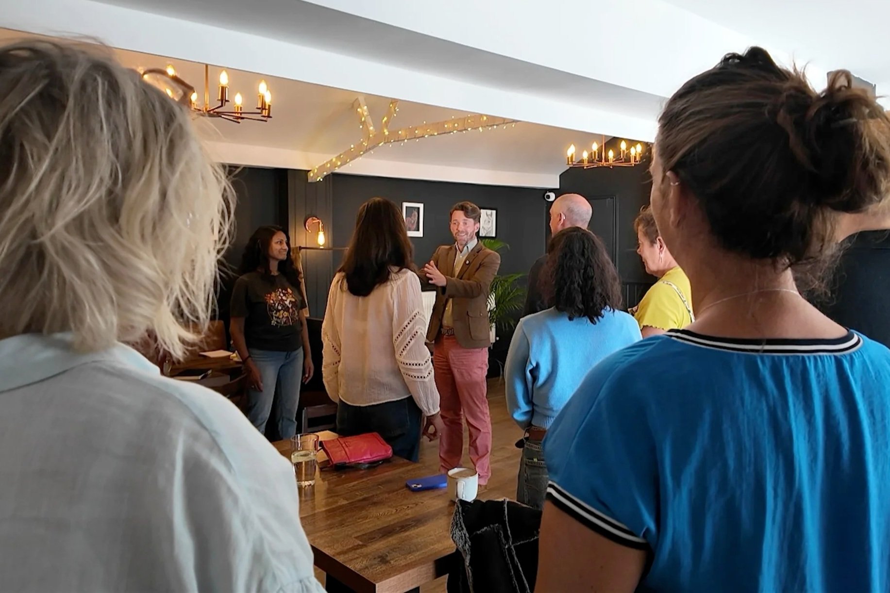 Group of people attending a meeting or gathering in a cozy, dimly lit room with black walls and warm lighting. The focus is on a man in a brown jacket speaking to the group, with others standing and listening around him.