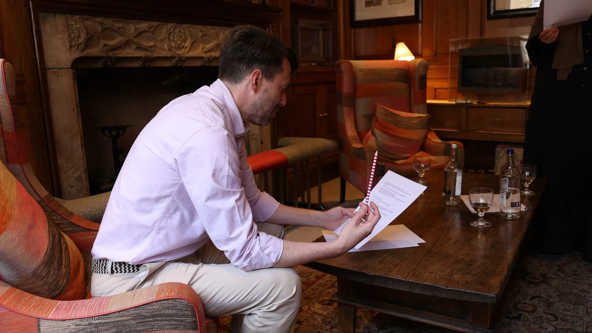 A man with dark hair and a beard, wearing a light pink shirt, is sitting at a wooden table, reading a piece of paper with a red and white striped pencil in his hand. The table has several glasses of water and a couple of bottles. The background features wooden paneling, framed art on the walls, a fireplace, and a lamp.