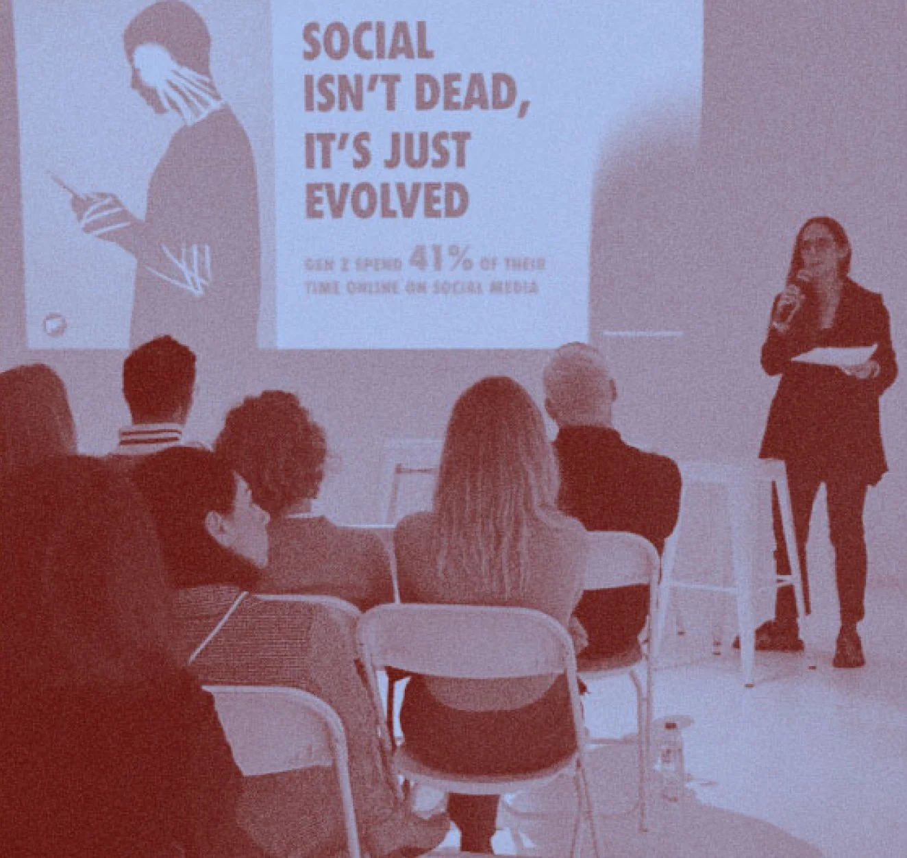 Photograph of Connie Flude holding a microphone and presenting to a seated audience, a projected slide reading "Social isn't dead, it's just evolved" visible behind her.  Duotone blue and grain effect applied.