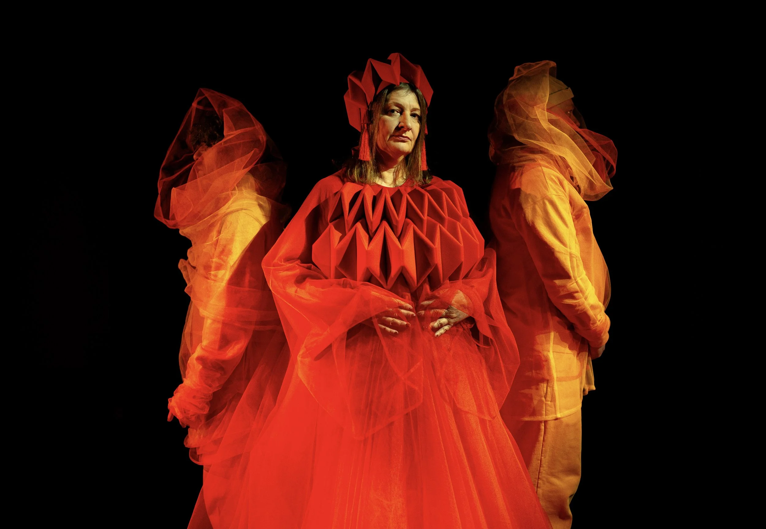 Woman in a red dress with 2 veiled characters behind her looking outwards.