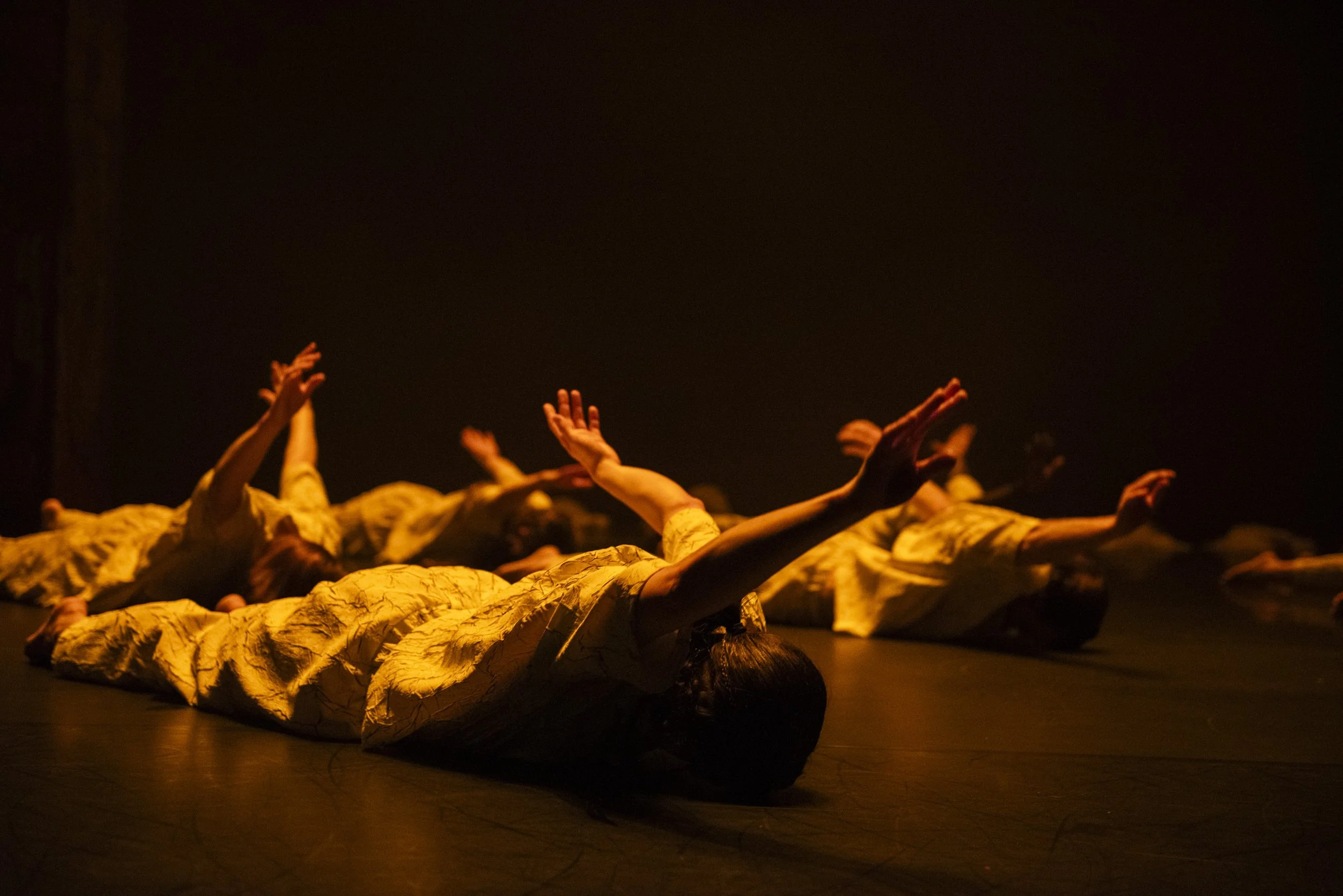 Dancers in white costumes lie on the floor with arms raised.