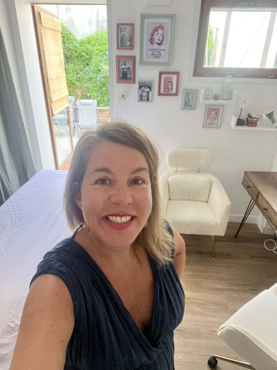Une femme souriante prend un selfie dans une pièce claire avec des murs blancs, décorée de plusieurs cadres photo et illustrations, y compris une affiche de style vintage, et une fenêtre laissant entrer la lumière naturelle.