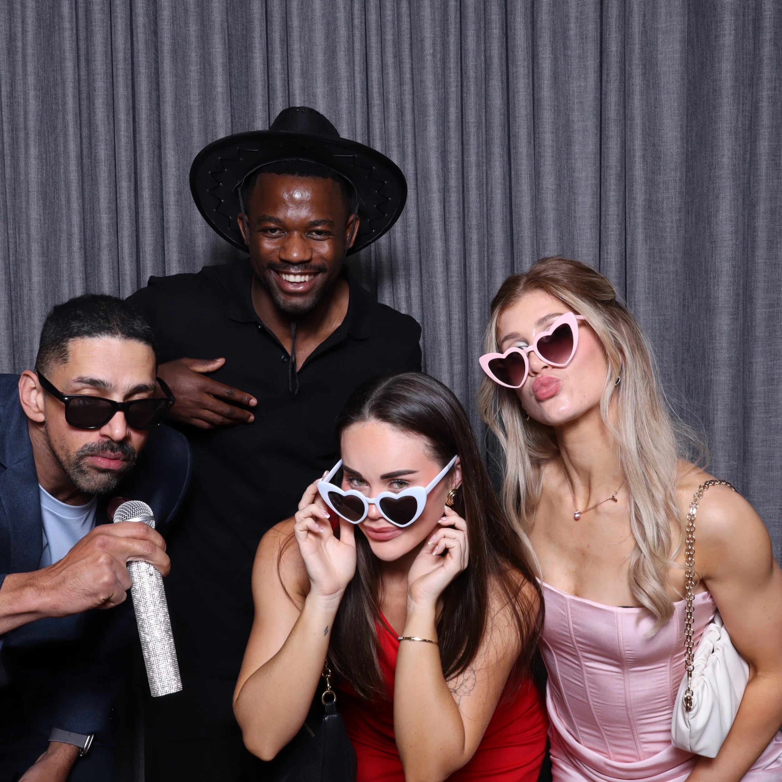 Group of four people dressed up with fun accessories, posing in front of a gray curtain. The group includes three women with sunglasses and a man wearing a hat.