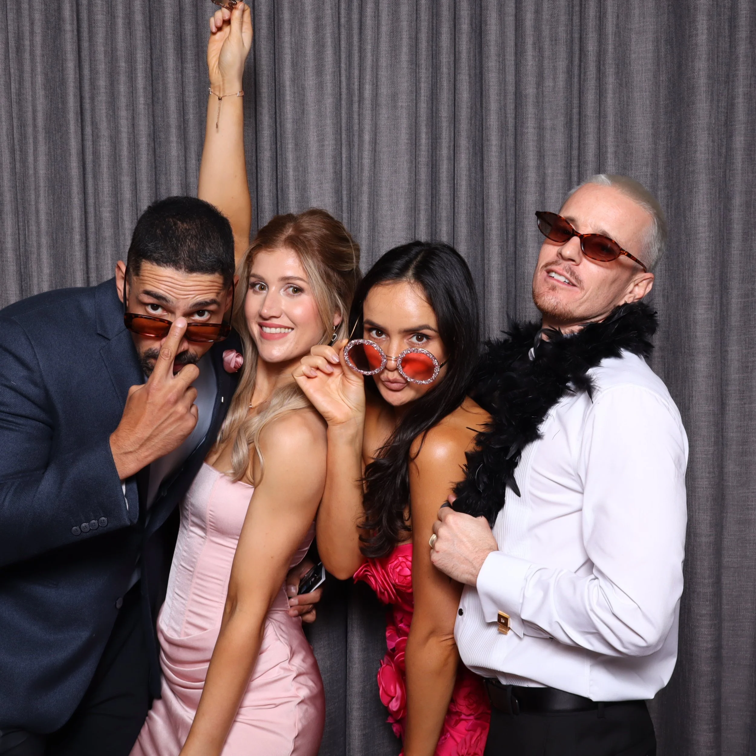 Four adults dressed up at a party, posing in front of a gray curtain. They are wearing accessories such as sunglasses, glasses, and a feather boa, and appear to be having fun.