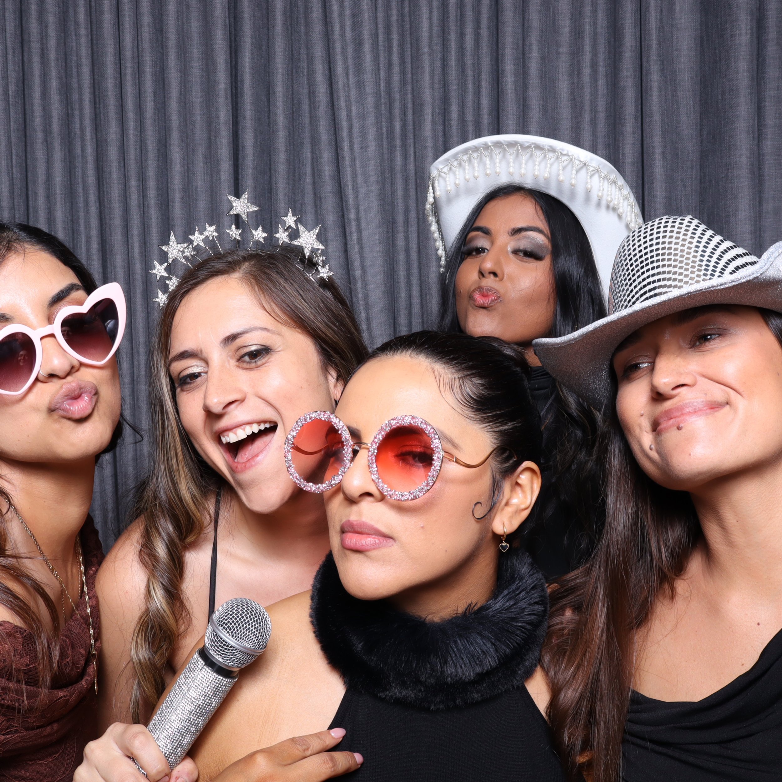 Five women dressed up with sunglasses and hats, posing for a photo booth at a party or celebration.