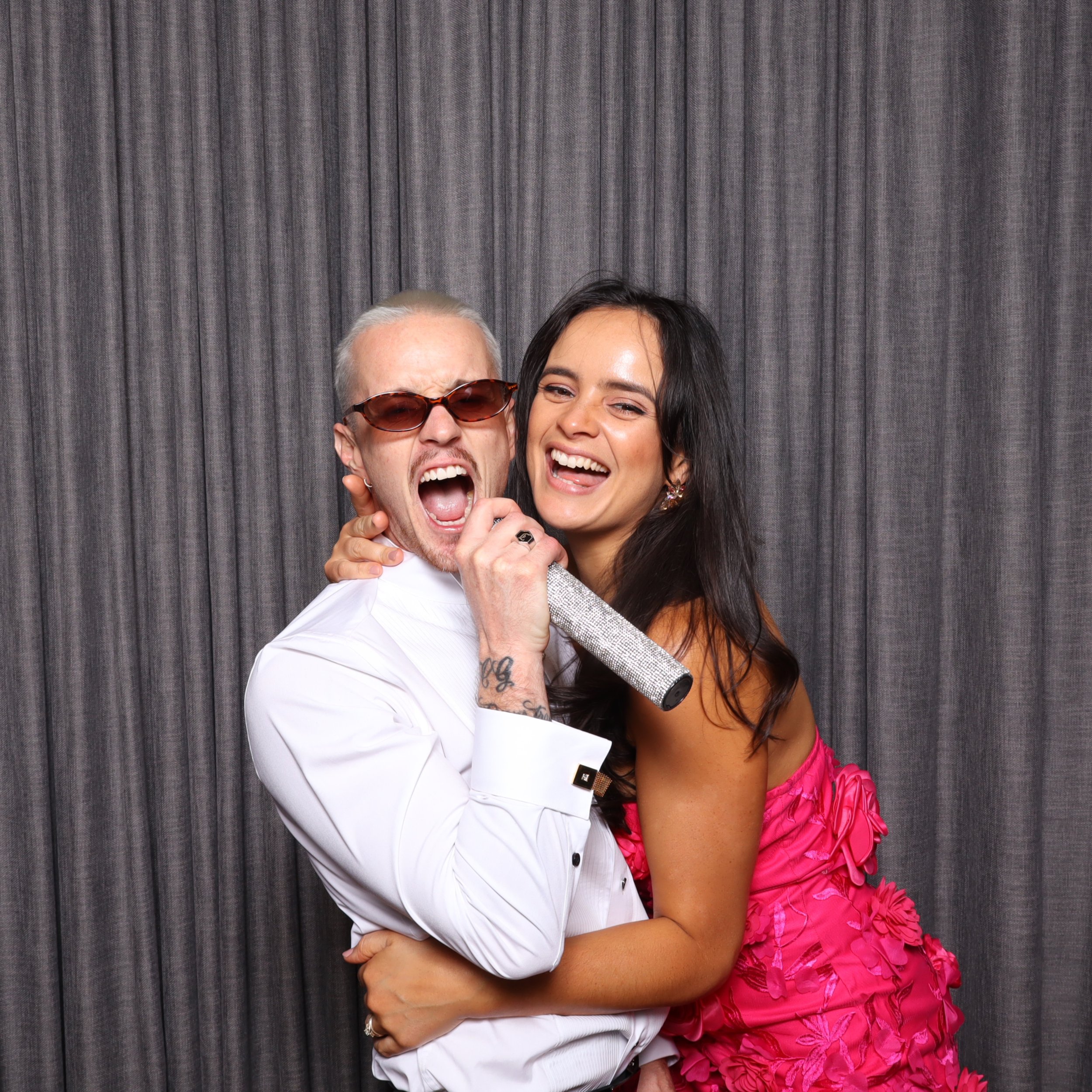 A man wearing sunglasses and a white shirt singing with a woman in a pink dress, both smiling and holding a microphone, against a gray curtain background.