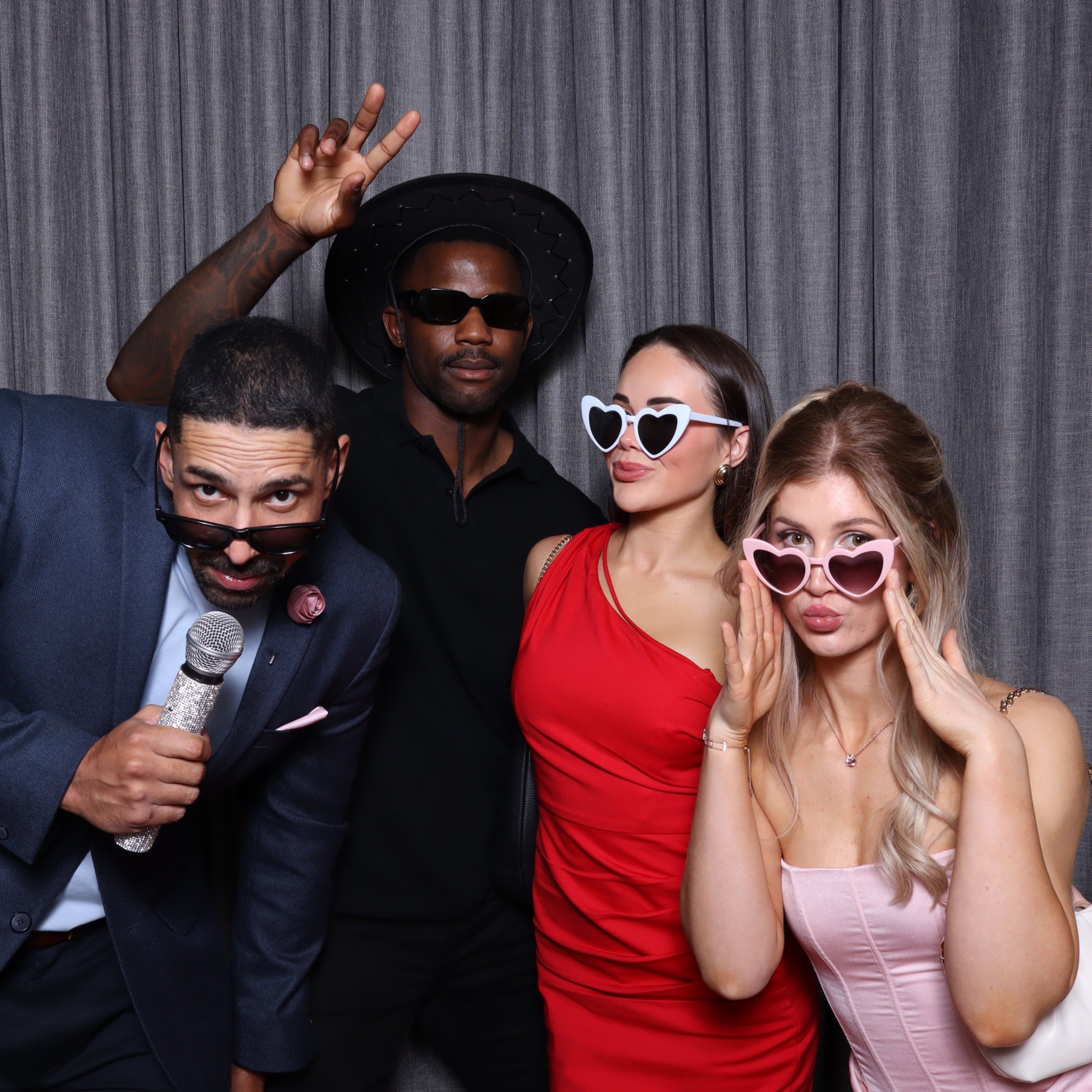 Four people posing at a party, wearing sunglasses and making playful expressions, with a curtain backdrop.