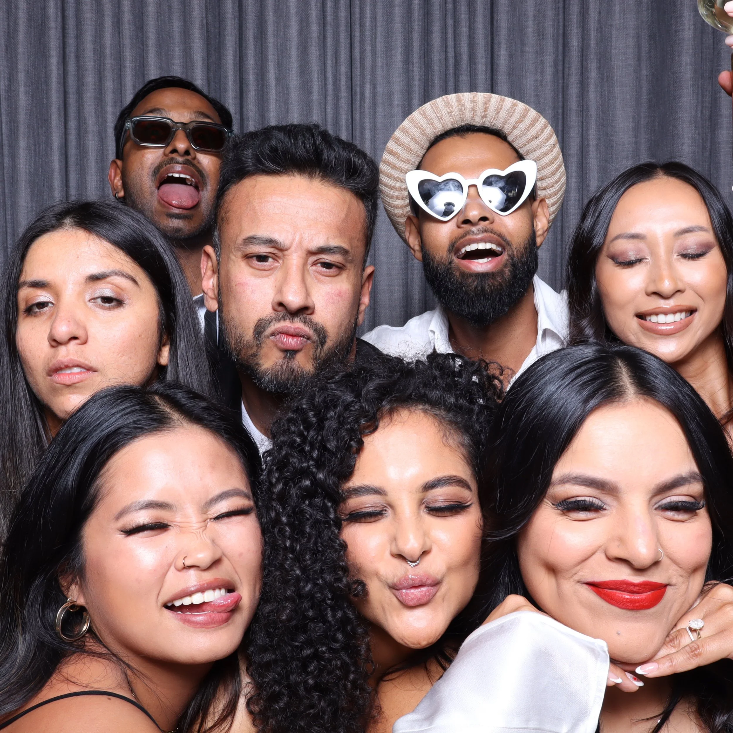 Group of nine diverse people posing together at a photo booth, wearing sunglasses and making playful faces, in front of a dark curtain background.