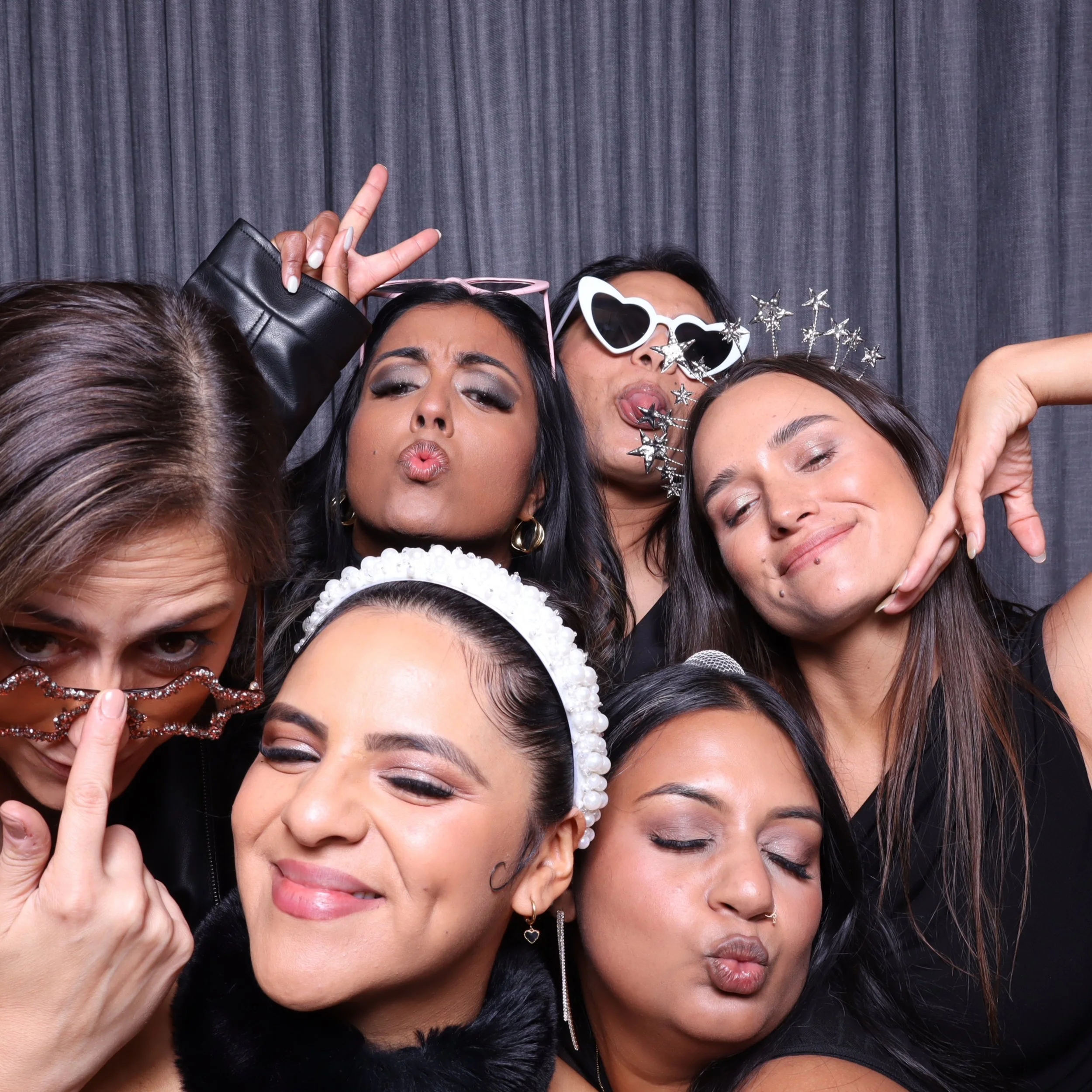 Seven women dressed for a party, making playful and silly faces with fun accessories, posing close together against a gray curtain backdrop.