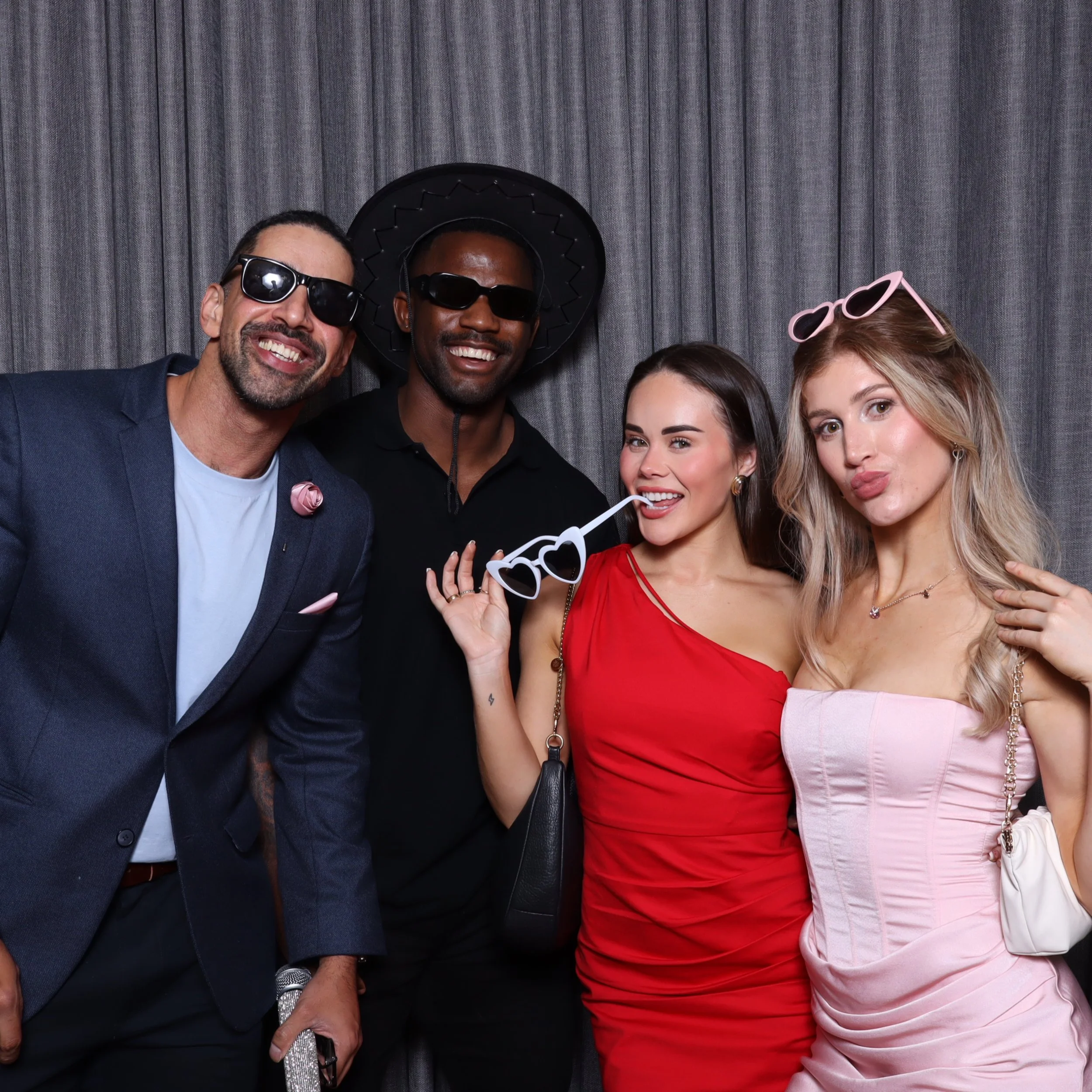 Four friends dressed up and wearing sunglasses, posing together at a party against a gray curtain background.