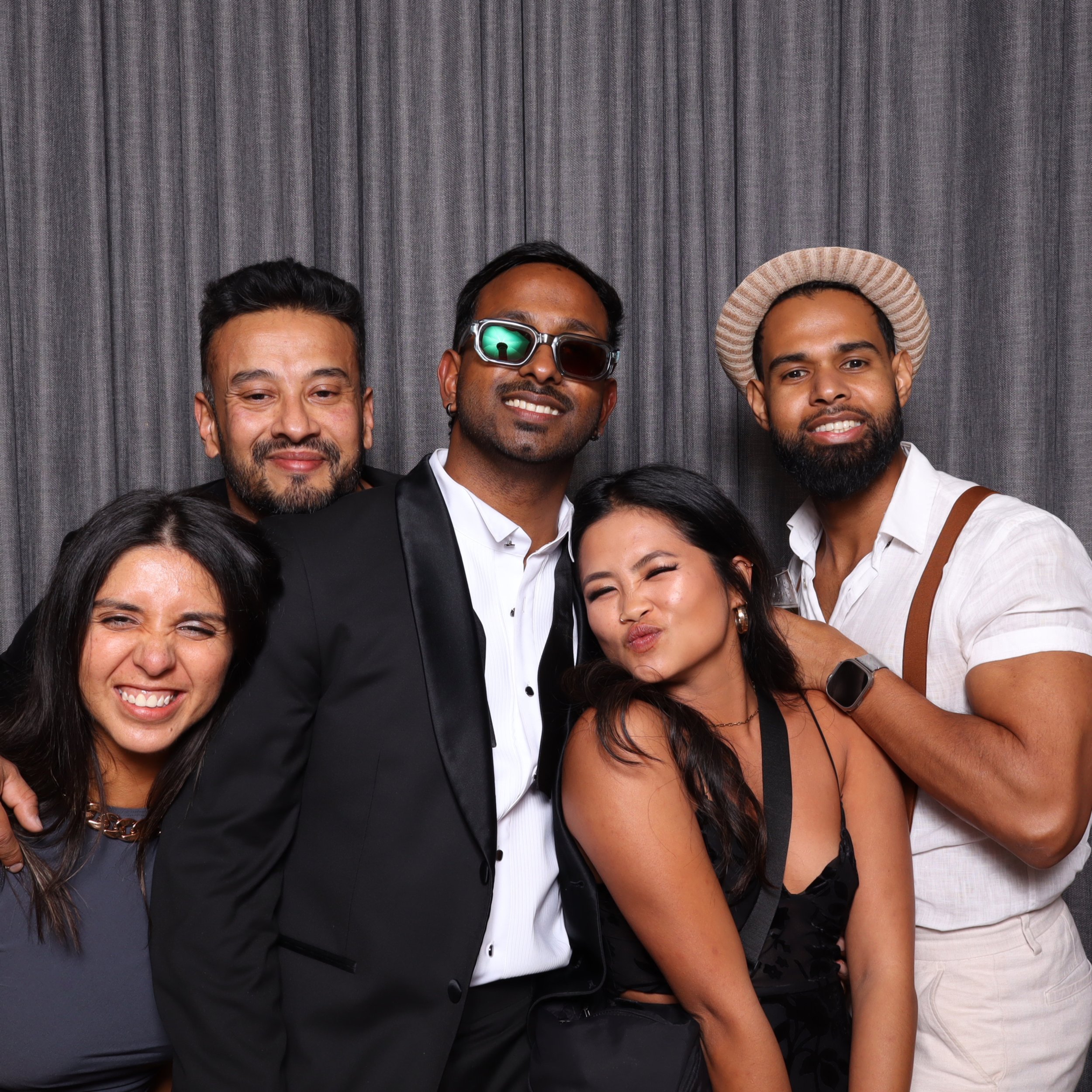Group of five diverse friends smiling and having fun at a photo booth, standing in front of a gray curtain backdrop.