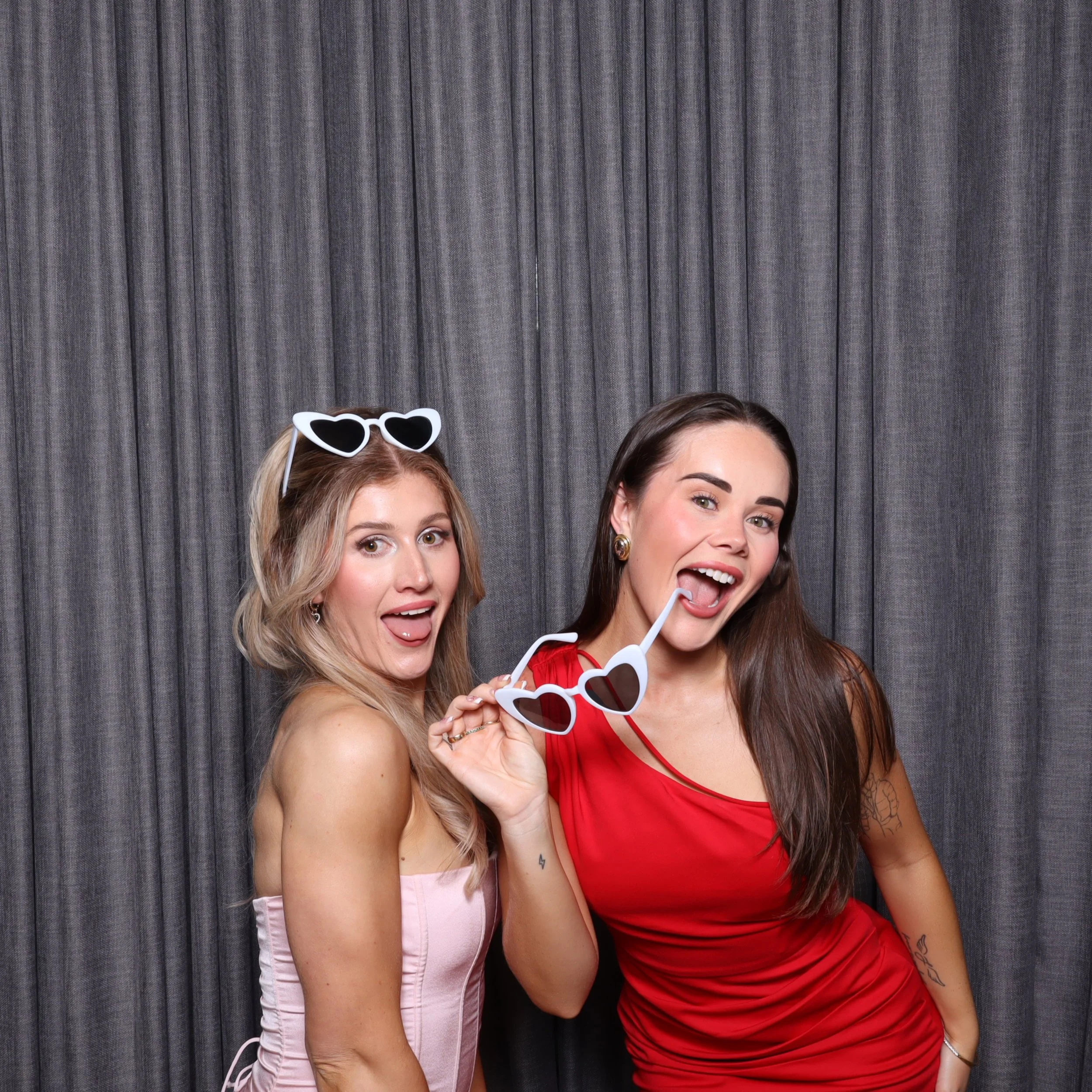 Two women posing in front of gray curtains, holding heart-shaped sunglasses, smiling and sticking out their tongues.