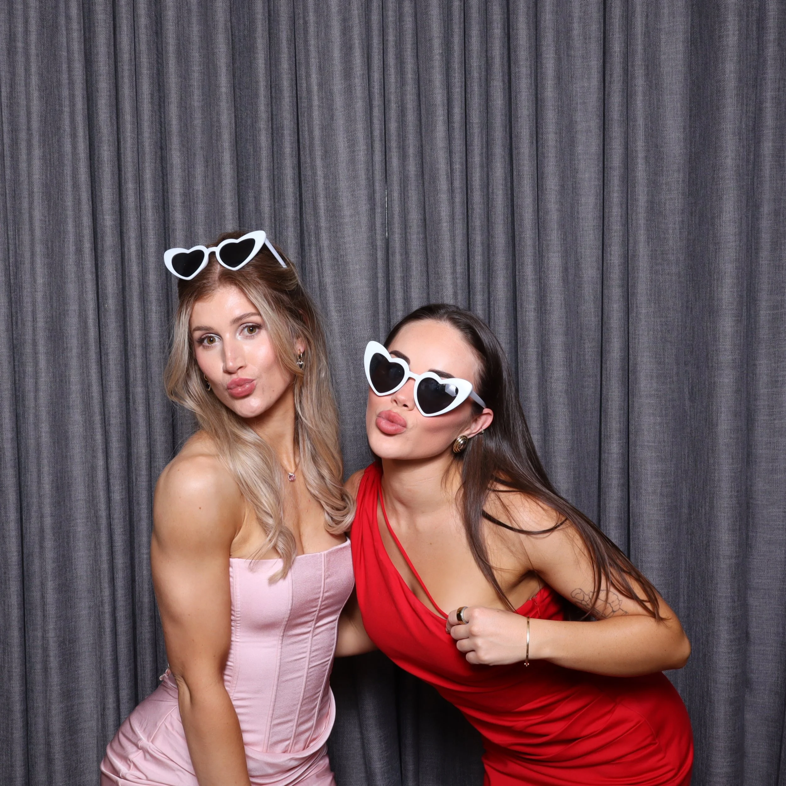 Two women wearing heart-shaped sunglasses, one in a pink strapless dress and the other in a red dress, posing playfully in front of a gray curtain.