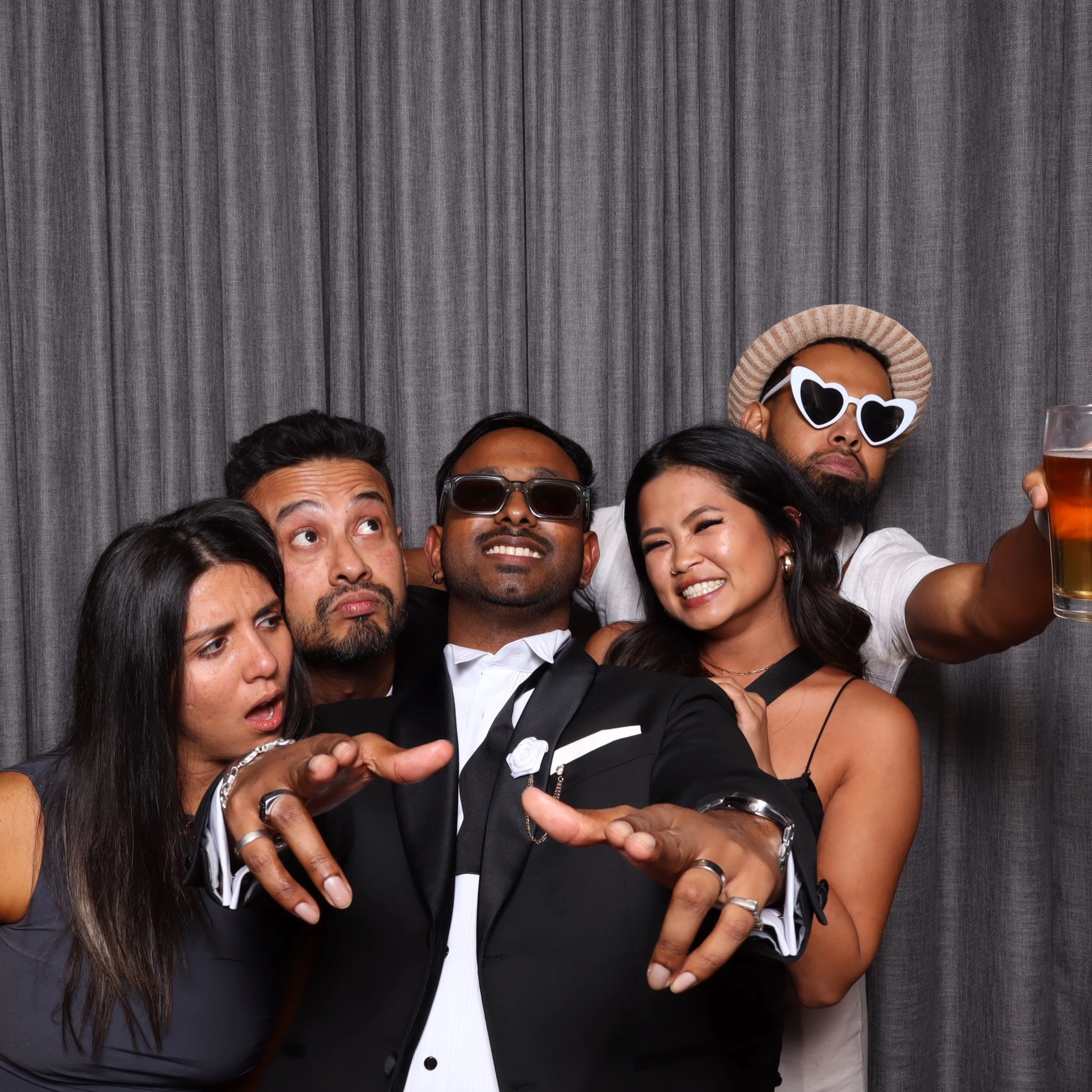 Group of five friends having fun at a photo booth wearing sunglasses and stylish outfits, with a gray curtain background.