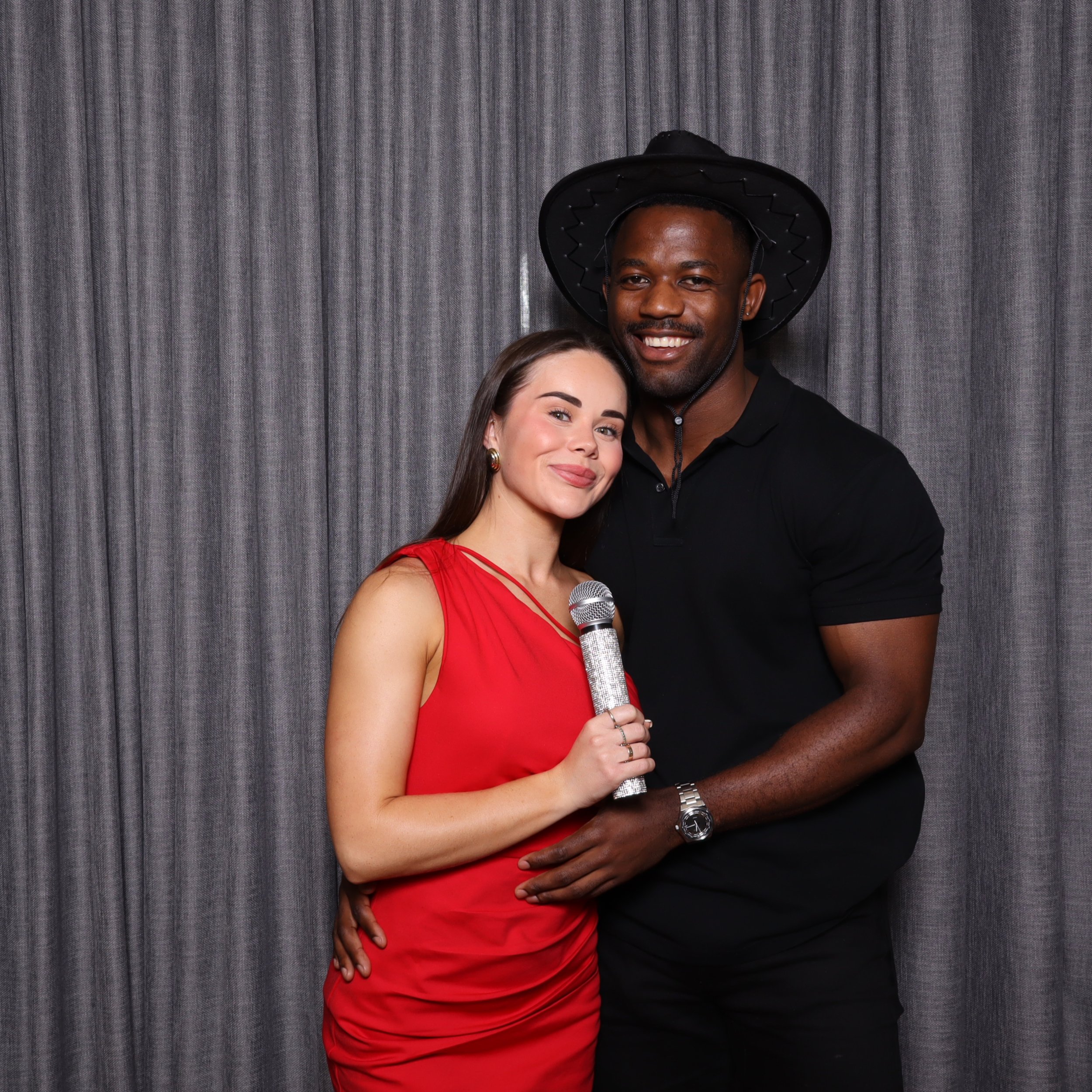 A woman in a red dress holding a microphone and a tall man in a black shirt wearing a large black hat standing together in front of gray curtains, smiling at the camera.