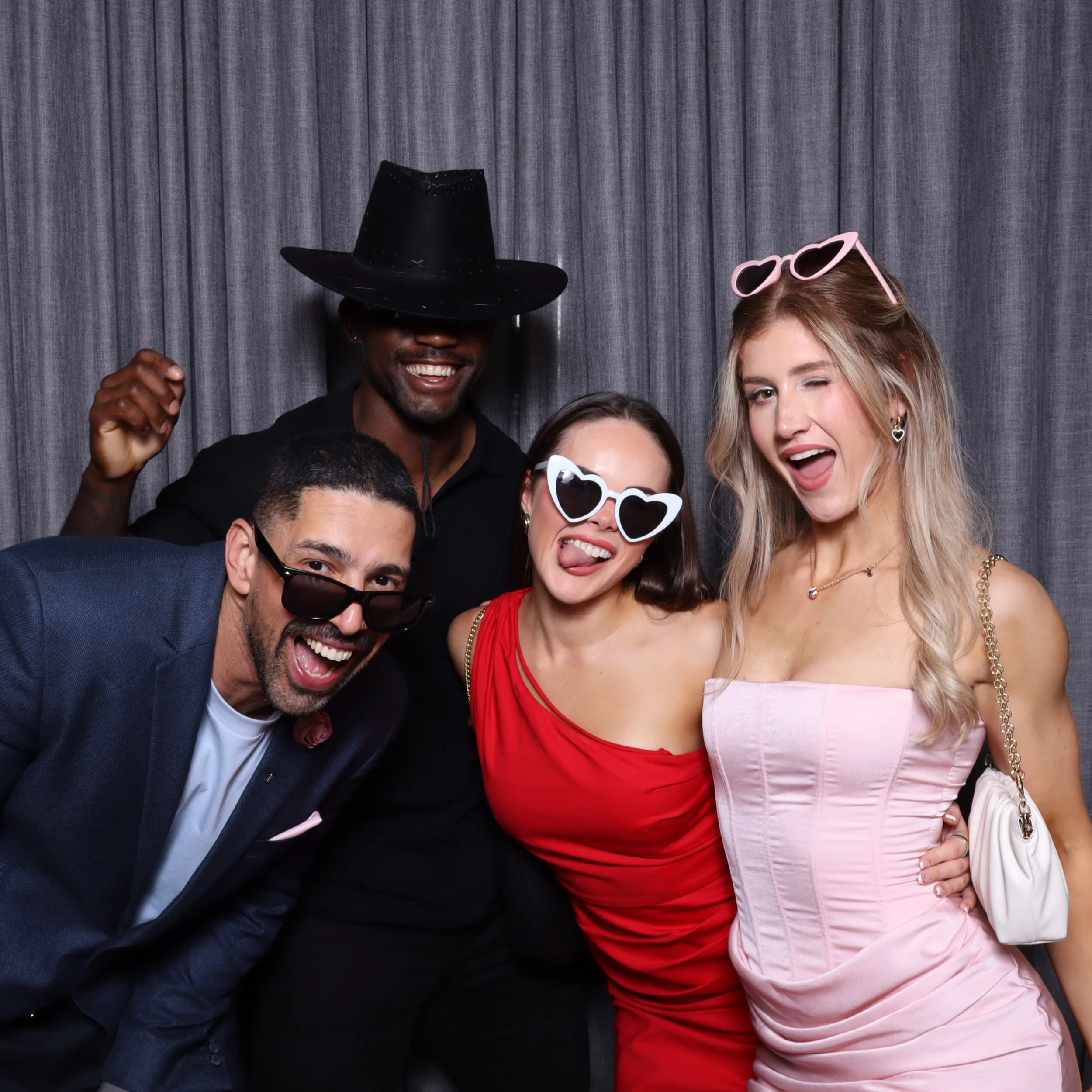 Group of four friends dressed up, wearing sunglasses and accessories, posing playfully in front of a gray curtain.
