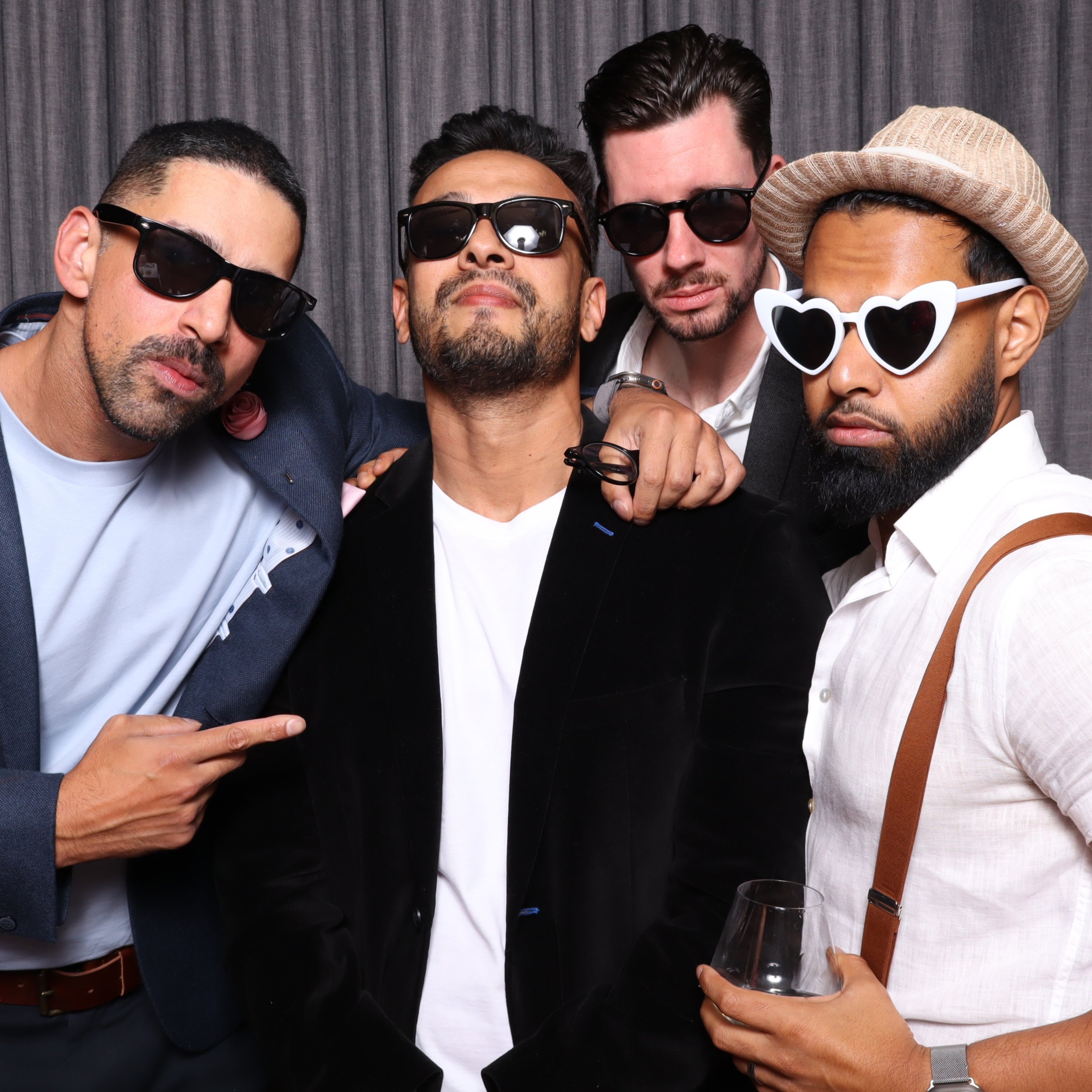 Five men posing together, wearing sunglasses, some with hats, in front of a gray curtain backdrop.