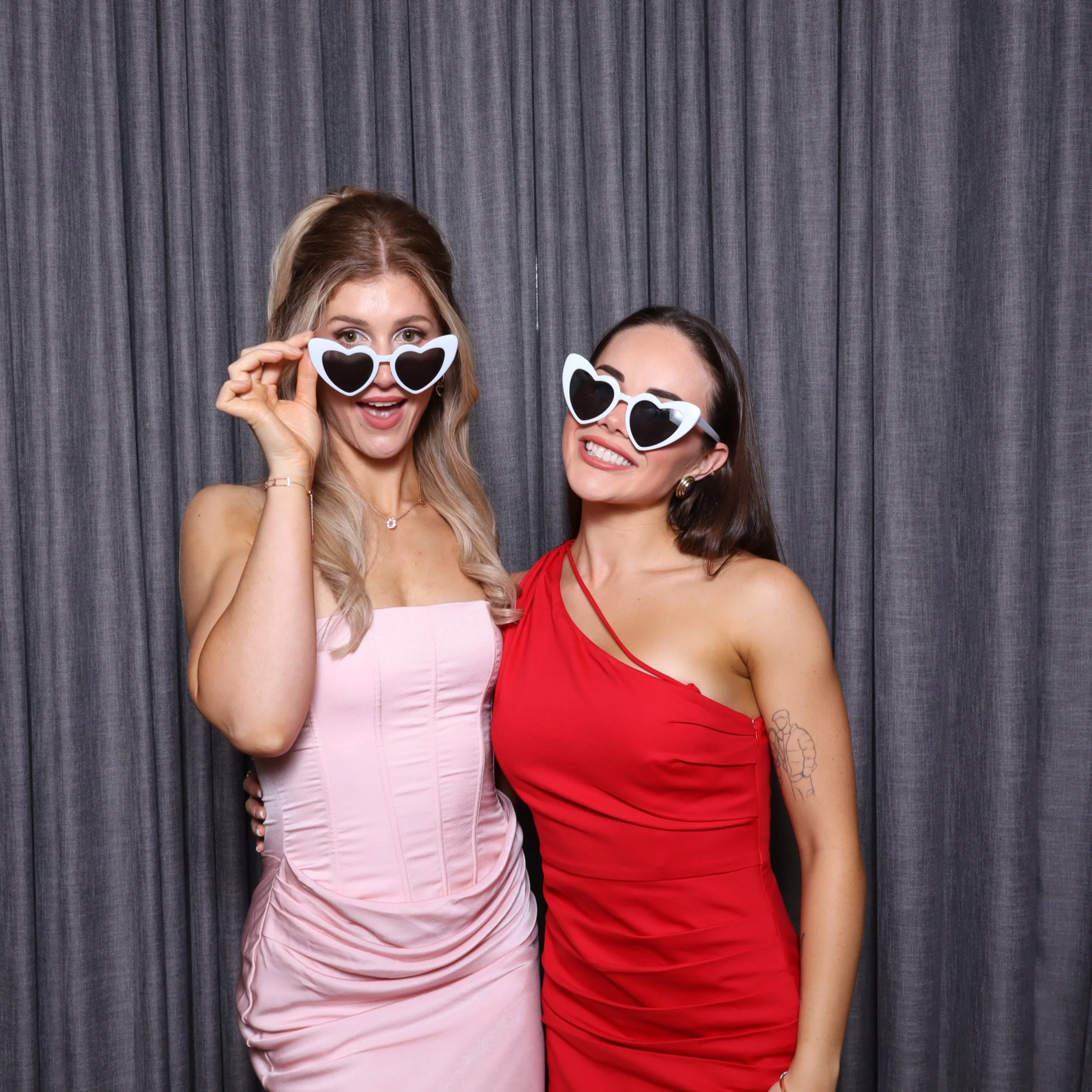Two women wearing heart-shaped sunglasses, smiling and posing in front of a gray curtain.