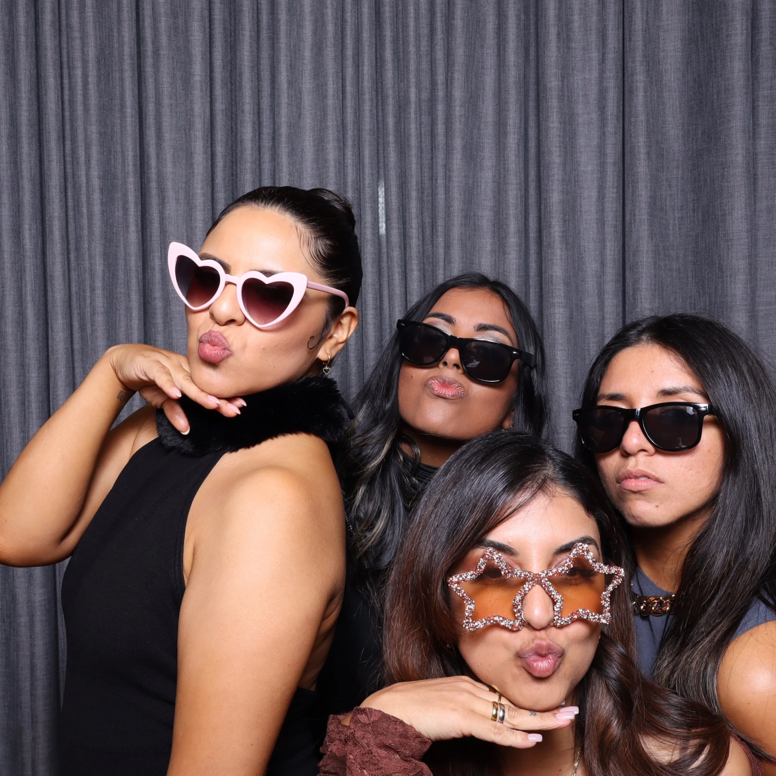 Four women posing with playful sunglasses and funky accessories in front of a gray backdrop.