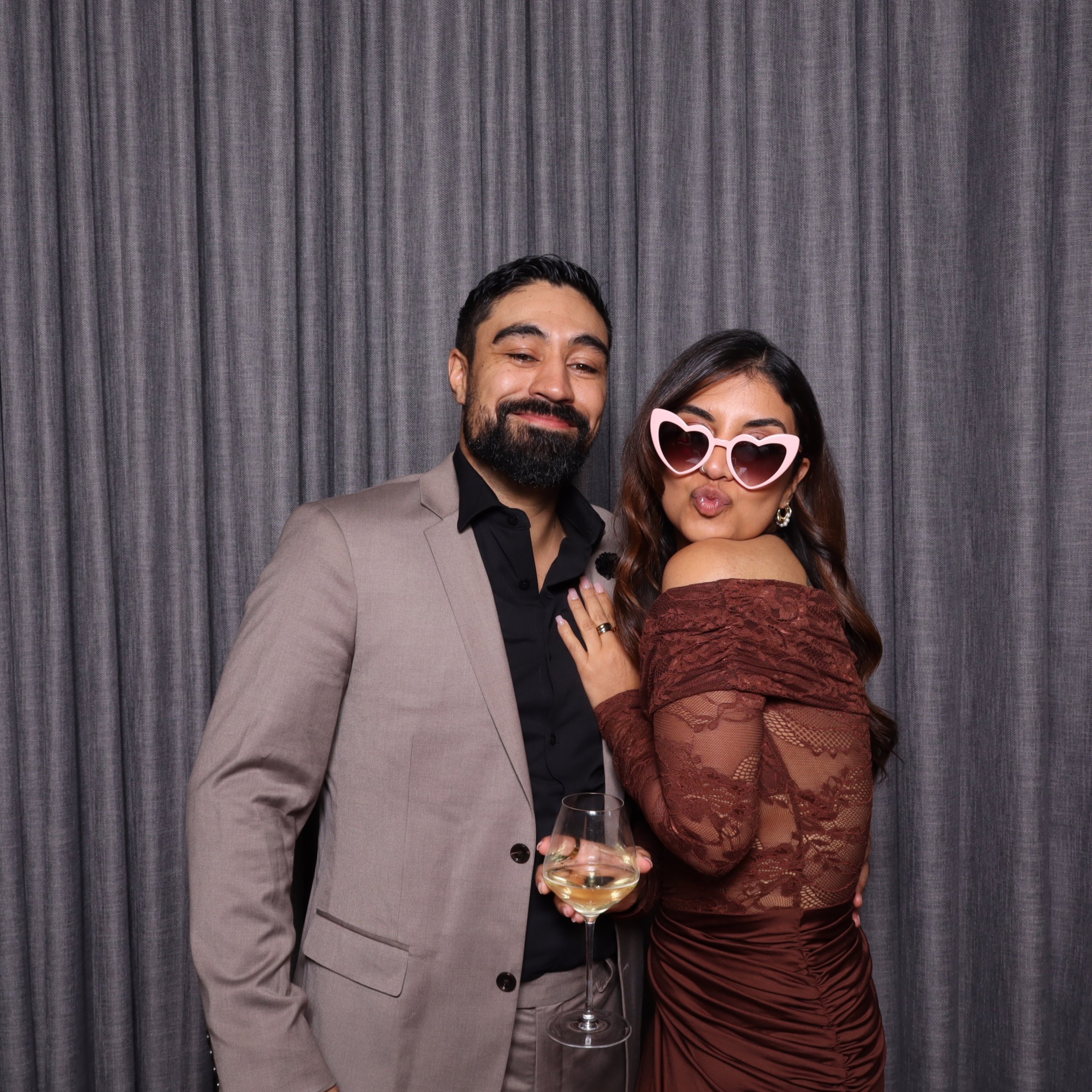 A man and woman dressed elegantly at a photo booth, with the woman wearing pink heart-shaped sunglasses and holding a glass of white wine, standing against a grey curtain background.