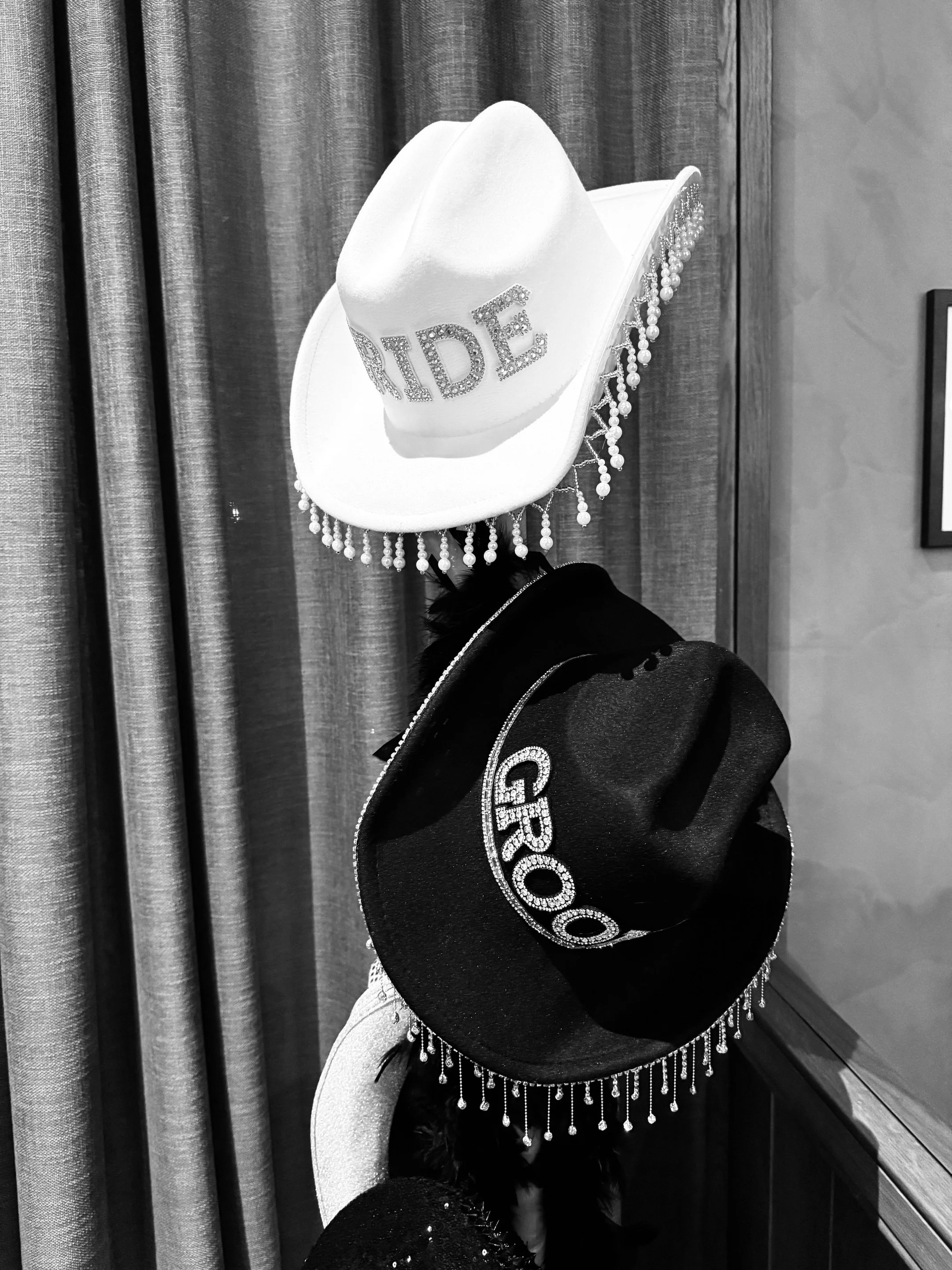 Two cowboy hats, one white with 'BRIDE' written on it and decorated with pearls, and one black with 'GROOM' written on it and decorated with pearls and feathers, hanging on a wall.