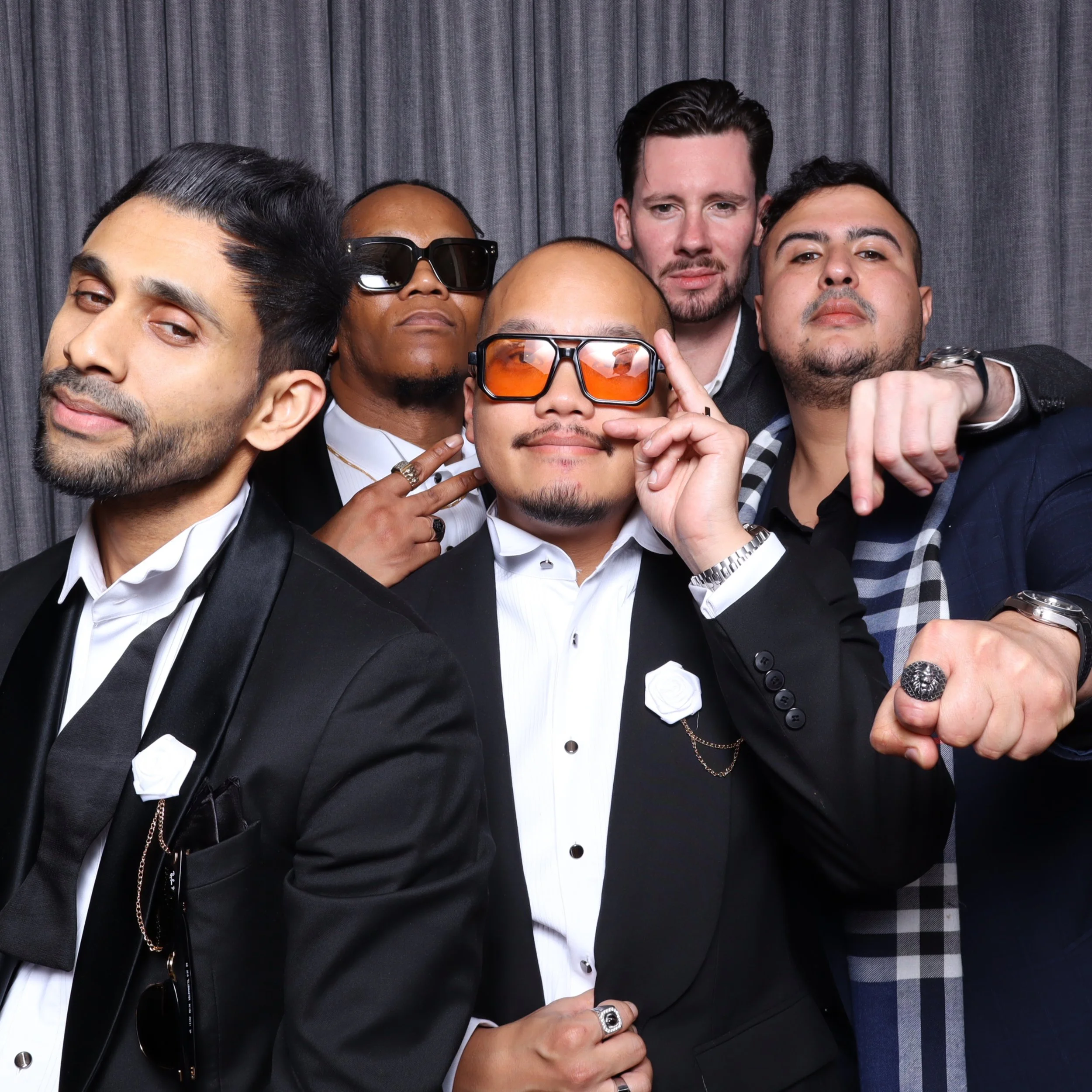 Group of five men dressed in tuxedos, posing confidently with some wearing sunglasses, at a formal event with a grey curtain backdrop.