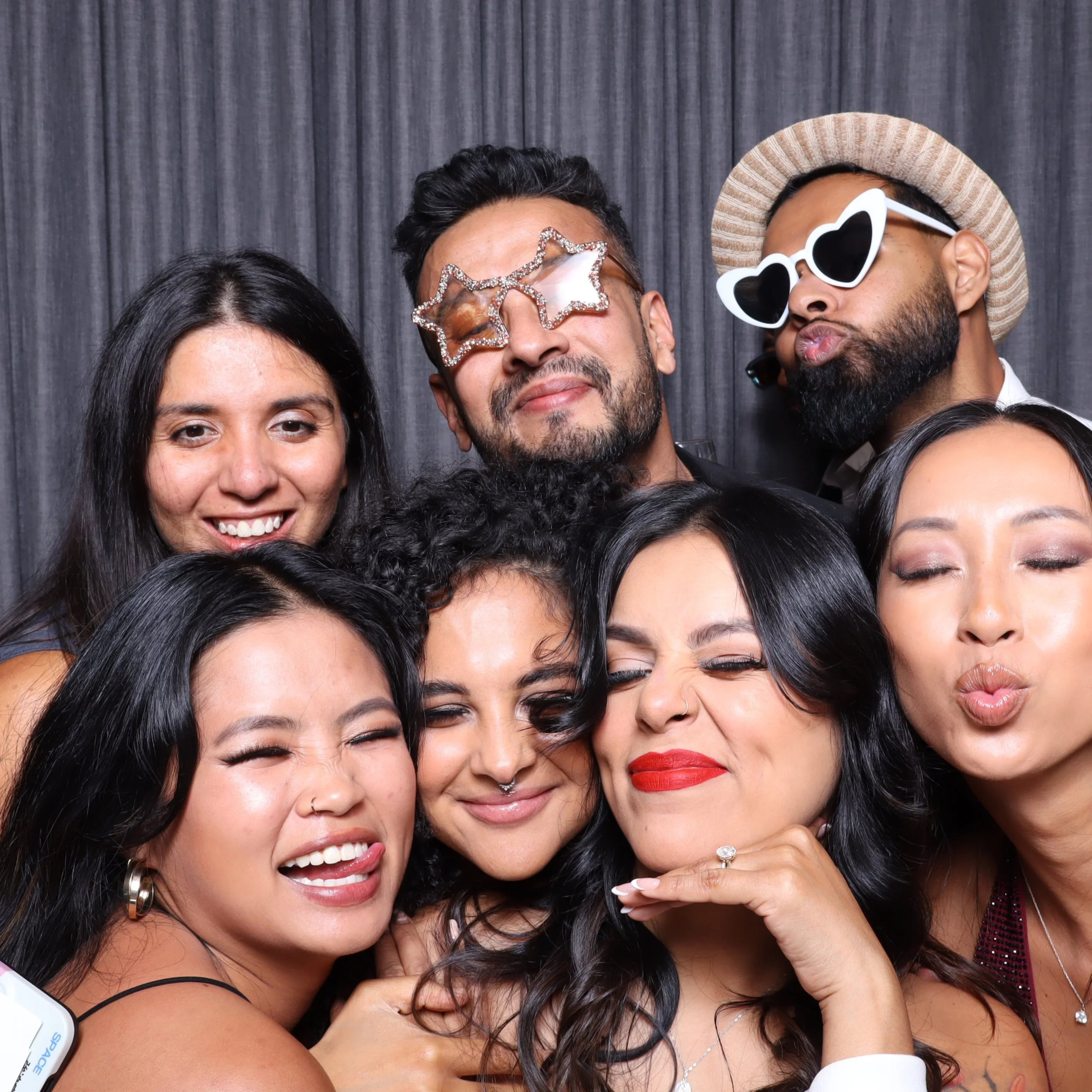 Group of seven diverse friends having fun at a photo booth, wearing playful accessories like star-shaped glasses, heart-shaped sunglasses, and a straw hat, making silly faces and smiling together.