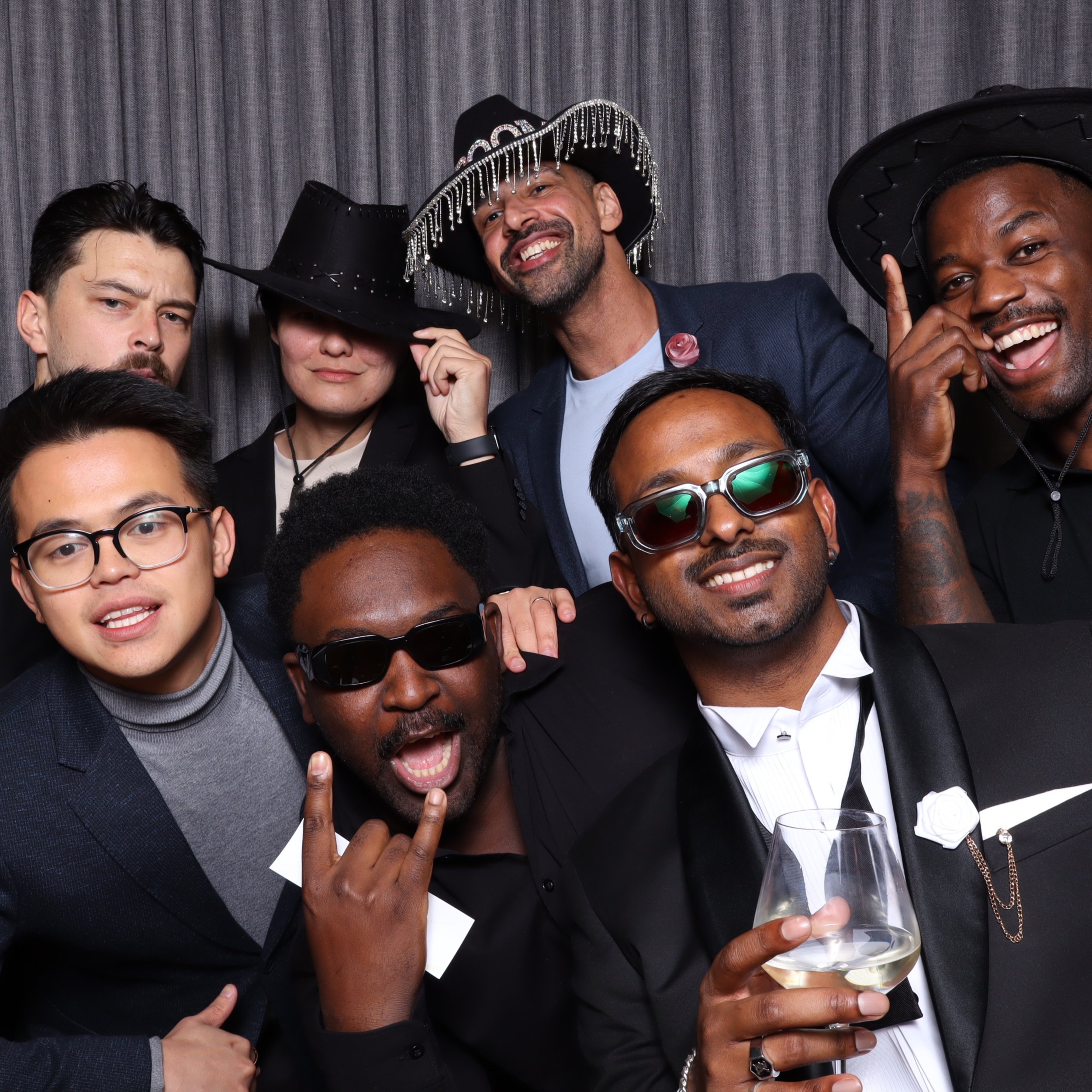 Group of diverse people at a celebration wearing tuxedos and fancy hats, smiling, with one person holding a glass of wine.