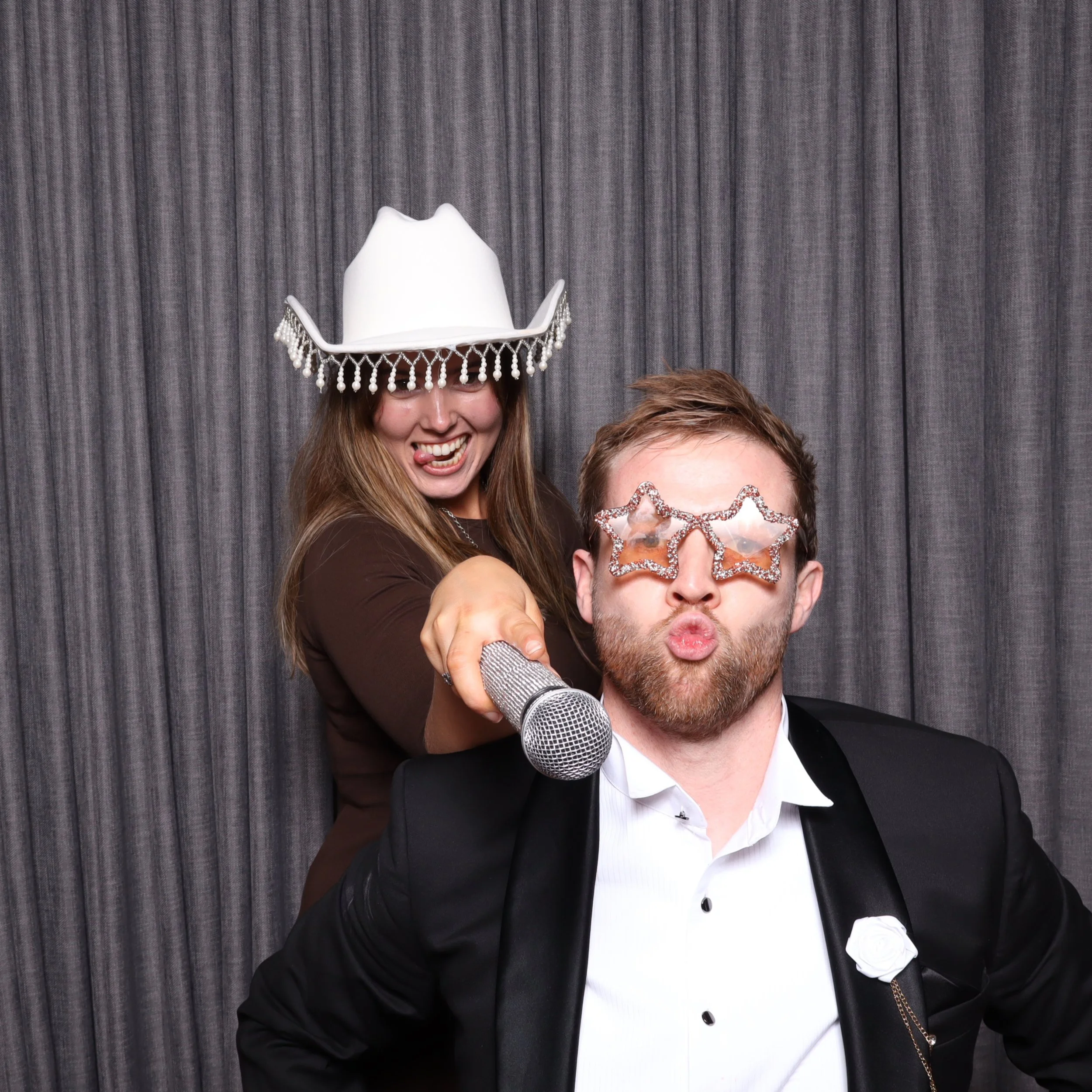 Two people wearing humorous accessories, with one holding a microphone. The woman is wearing a white cowboy hat with beaded fringe and the man is wearing star-shaped sparkly glasses, making kissing faces in front of a gray curtain background.