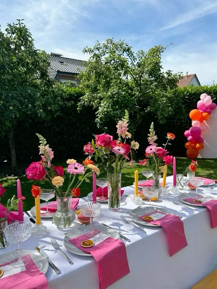 Table extérieure décorée avec des fleurs colorées, des bougies roses et orange, serviettes roses, verres à pied, et assiettes avec rondelles d'orange, dans un jardin avec des arbres et des ballons en arrière-plan.