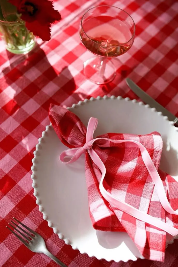 Table dressée avec assiette blanche, serviette rouge à carreaux nouée, verre de vin rosé et couverts sur nappe à carreaux.