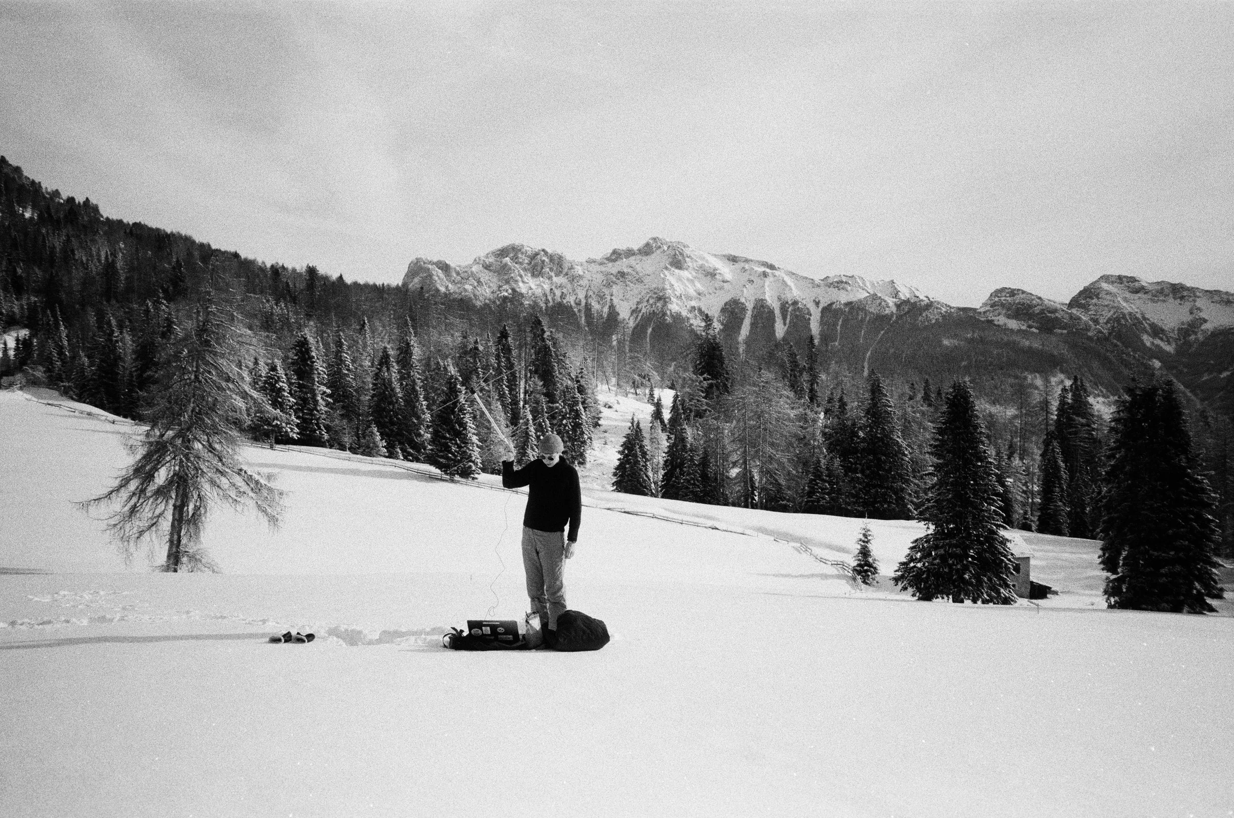 Person in winter clothing standing in snowy landscape with mountains and trees in the background, carrying equipment.