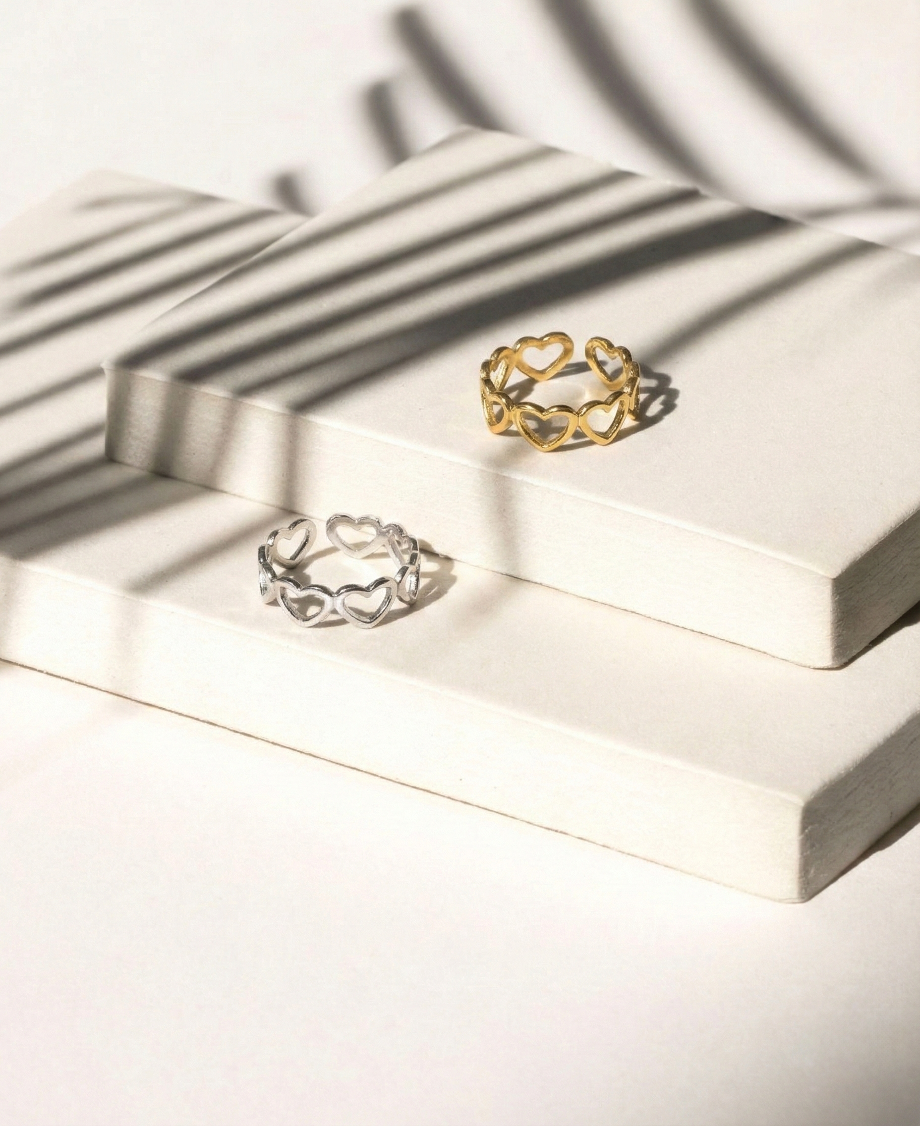 Zwei Ringe, einer in Silber und einer in Gold, mit Herzdesign, auf weißen Podesten mit Schatten von Palmenblättern